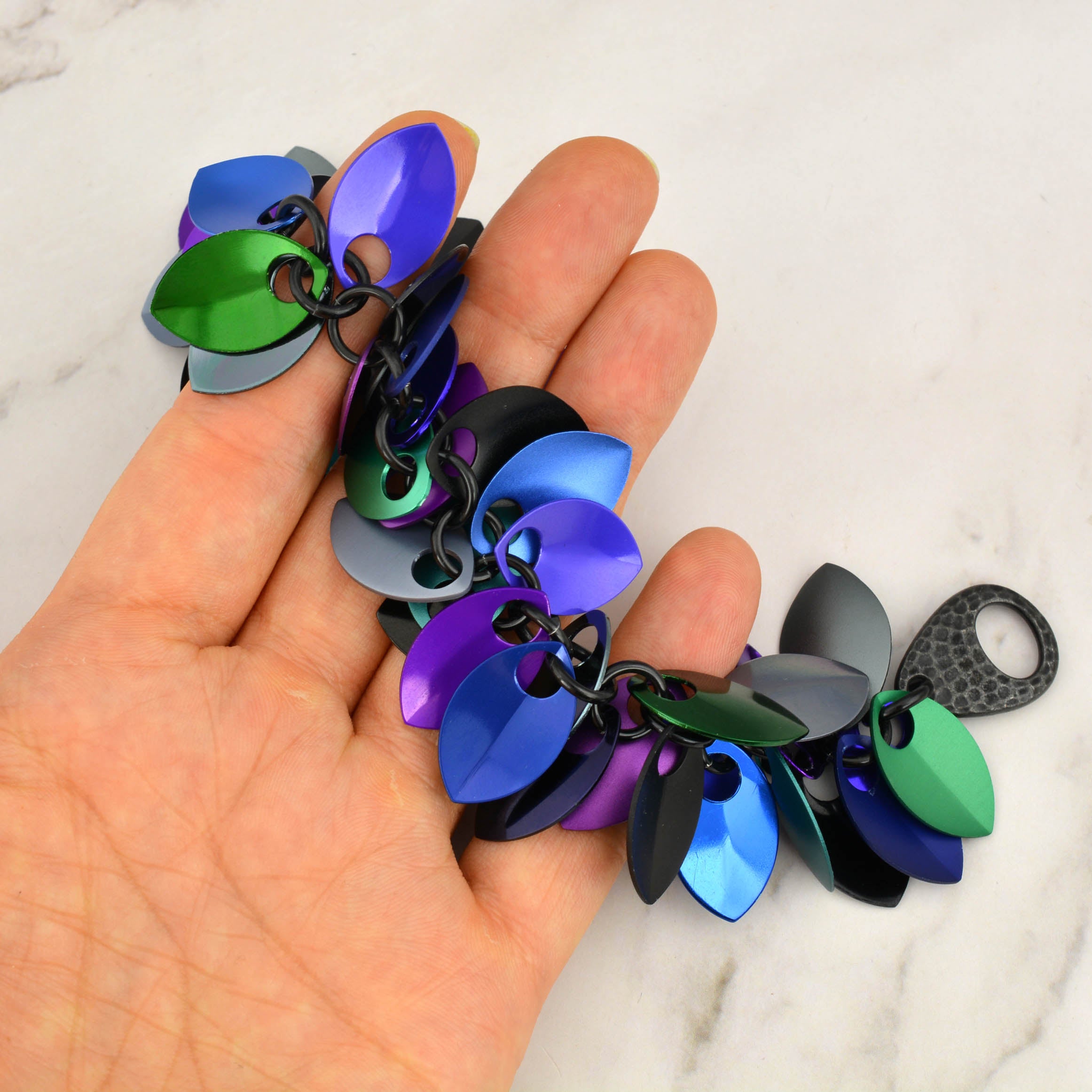 Dark Mermaid Cascading Leaves scalemaille bracelet (blue, purple, teal, black, green) held loosely in artisan Rebeca Mojica's hand, demonstrating the fluid movement and iridescent finish.
