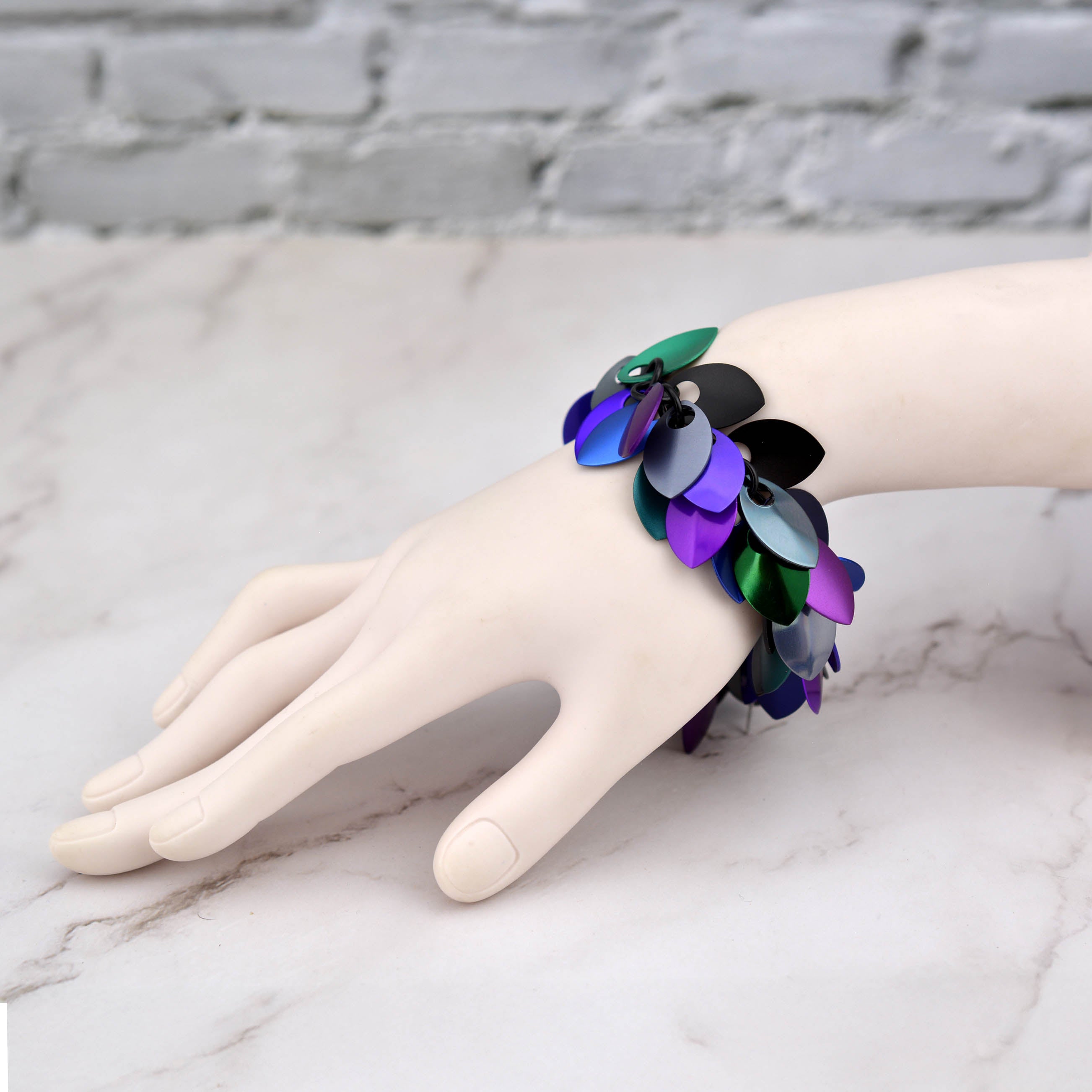 The Dark Mermaid scalemaille bracelet modeled on a white mannequin wrist, shown from a side angle with a marble and blurred white brick background.