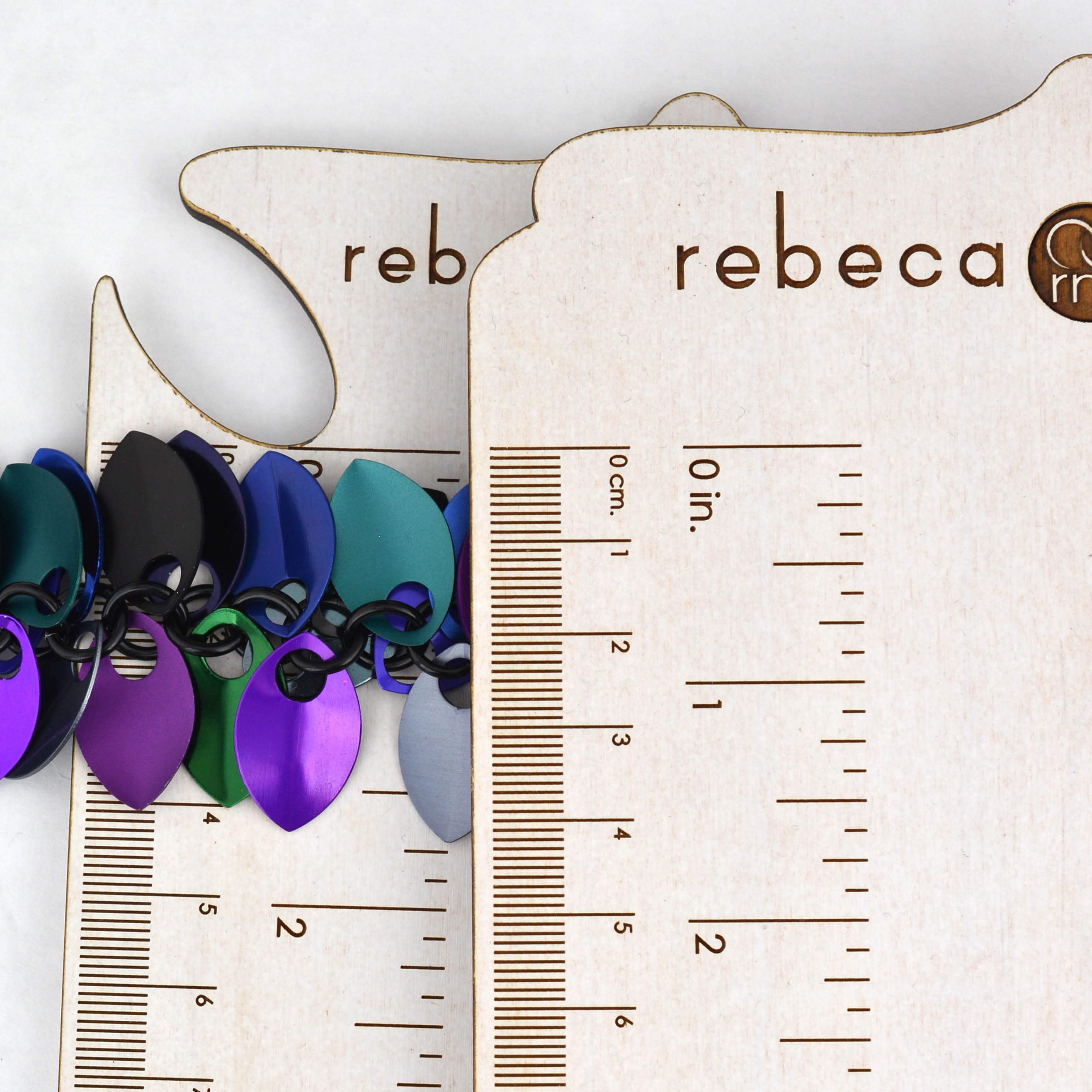 Extreme close-up of the anodized aluminum scalemaille links of the bracelet shown next to a wooden ruler for precise size reference in inches and centimeters. With the scales fully splayed, the bracelet measures about 1.5" across.