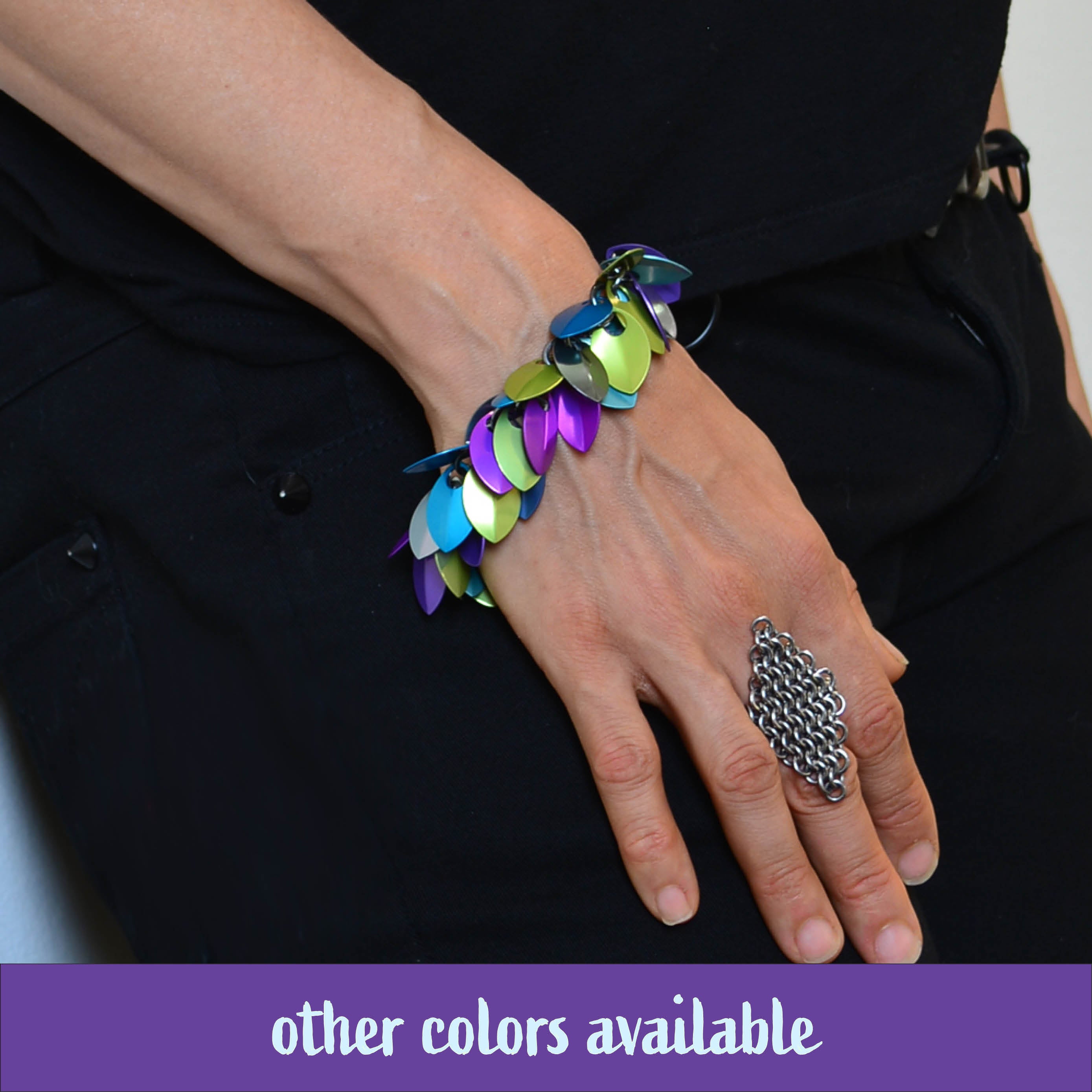 Close-up of artisan Rebeca Mojica's hand and wrist modeling the Cascading Leaves Bracelet in the Ocean Mermaid colorway (muted lime green, bright purple, and blue scales) against dark clothing. A banner reads, "other colors available.