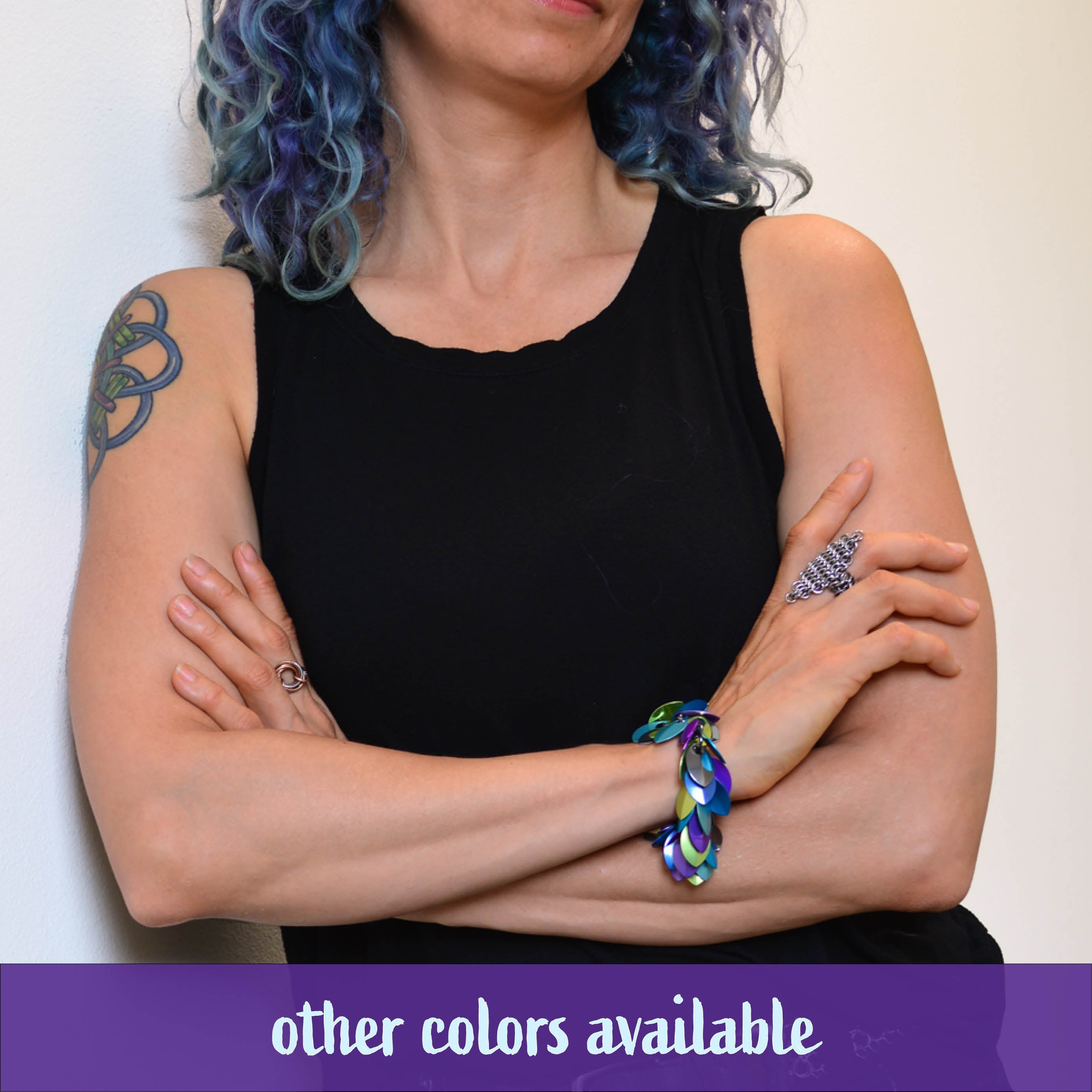 Artisan Rebeca Mojica with blue and purple curly hair and a tattoo on her arm wears a black tank top, with her arms crossed. She is modeling the Cascading Leaves Bracelet in a mix of muted lime green, purple, blue, and teal on her right wrist. A banner reads, "other colors available."