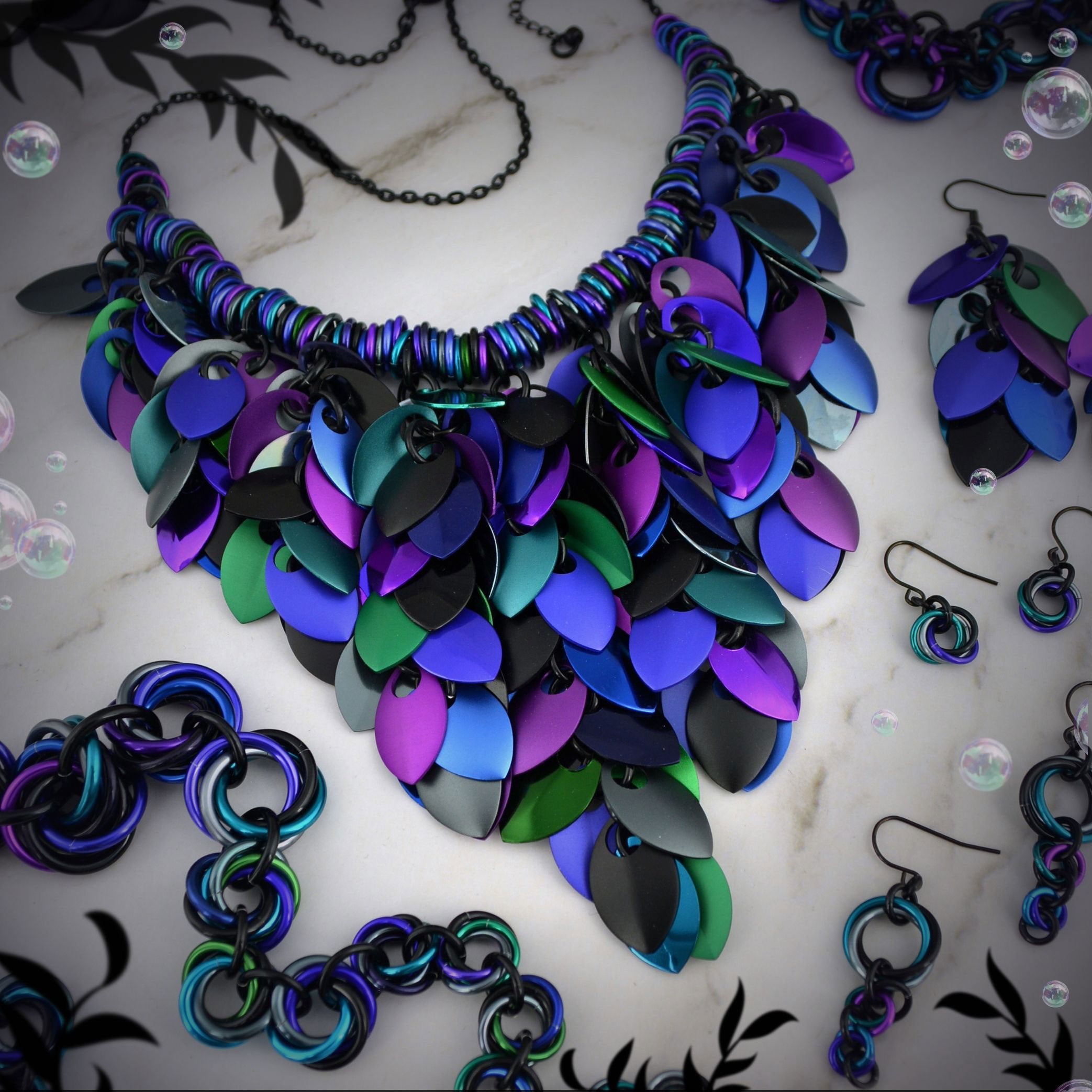 Dark Mermaid chainmail jewelry collection flat lay by Rebeca Mojica: Feathered Bib Statement Necklace, Feathered Earrings, Single Knot Earrings, and bracelets in iridescent teal, blue, purple, and black.