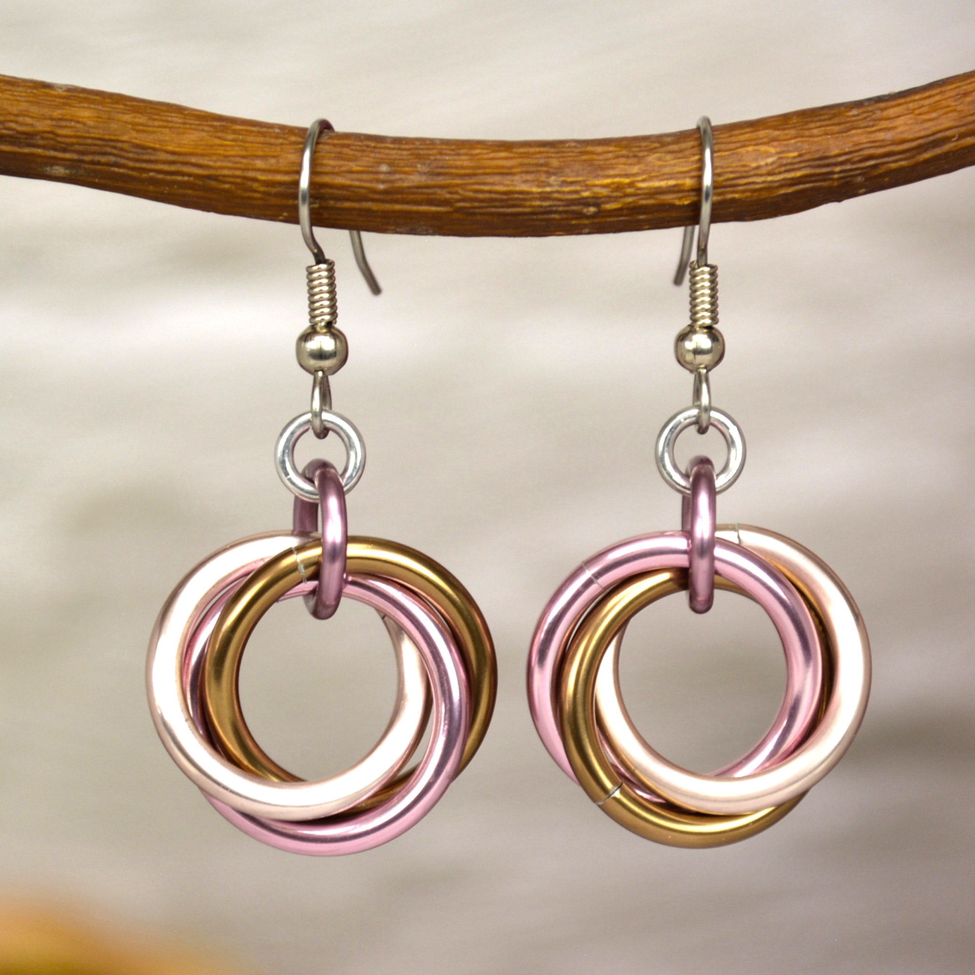 Soft pink and brown chainmaille earrings hanging from a branch. Each earring has a "knot" of 3 large jump rings in rose gold, russet and pink on a stainless steel earwire. The background is a blurry off-white color brick wall.