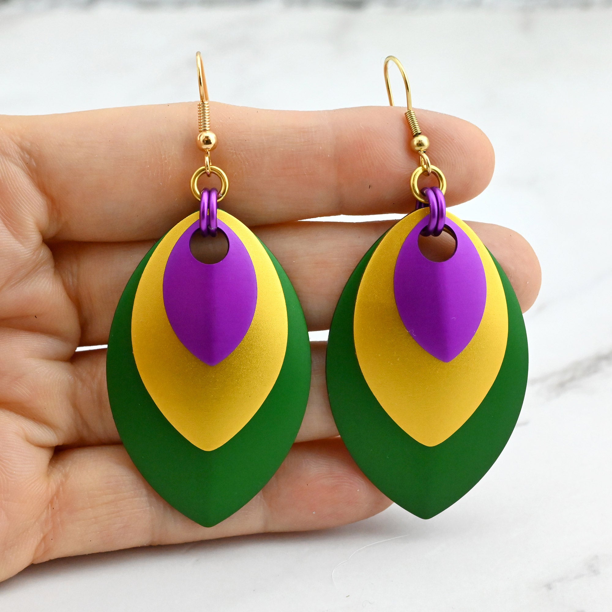 Stacked leaf earrings in purple, gold, and green held against fingertips to demonstrate the large leaf-shaped components.