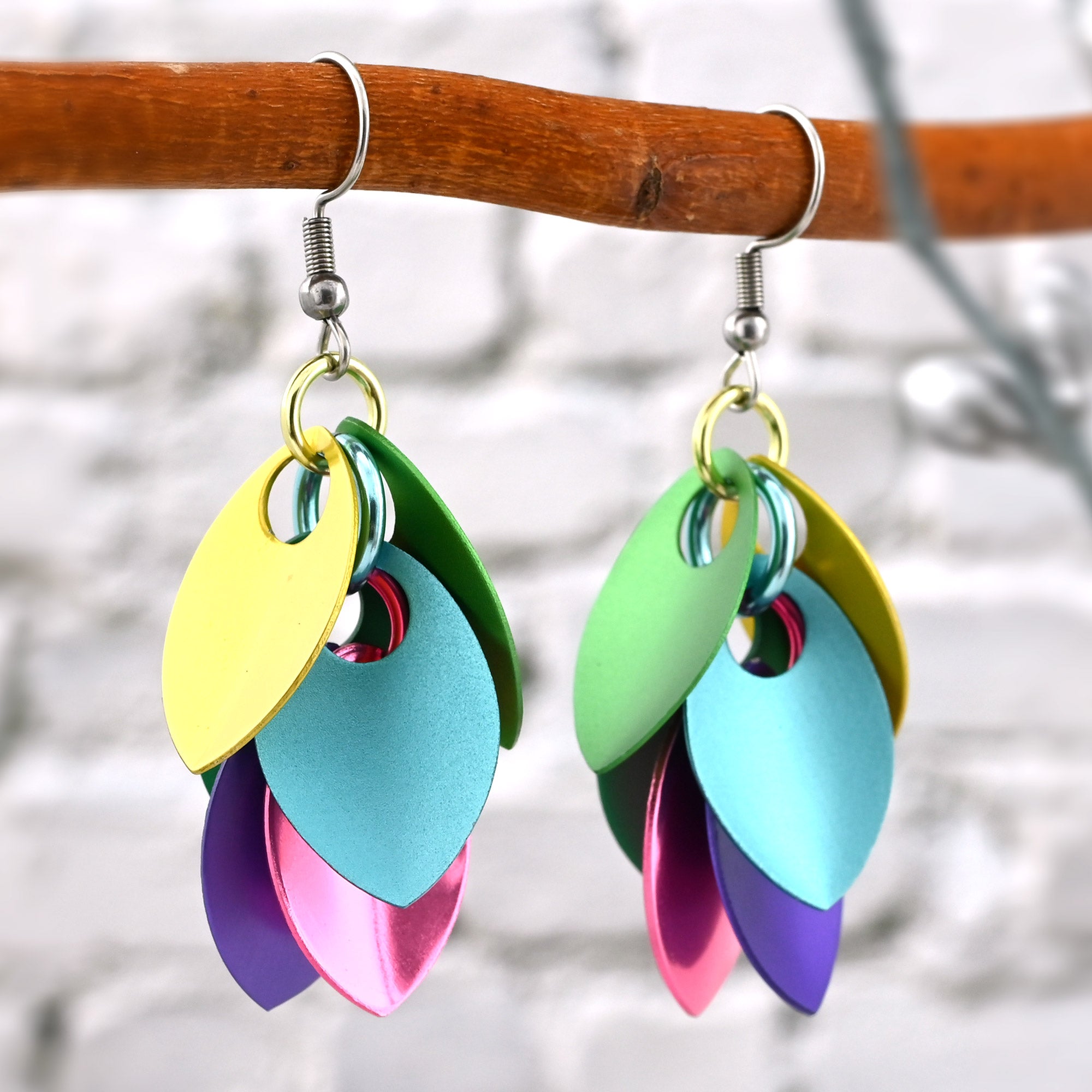 A pair of multi-color Cascading Leaves earrings hanging from a branch against a white brick background. The aluminum scales in the earrings are a variety of saturated pastels including yellow, blue, green, pink and purple.