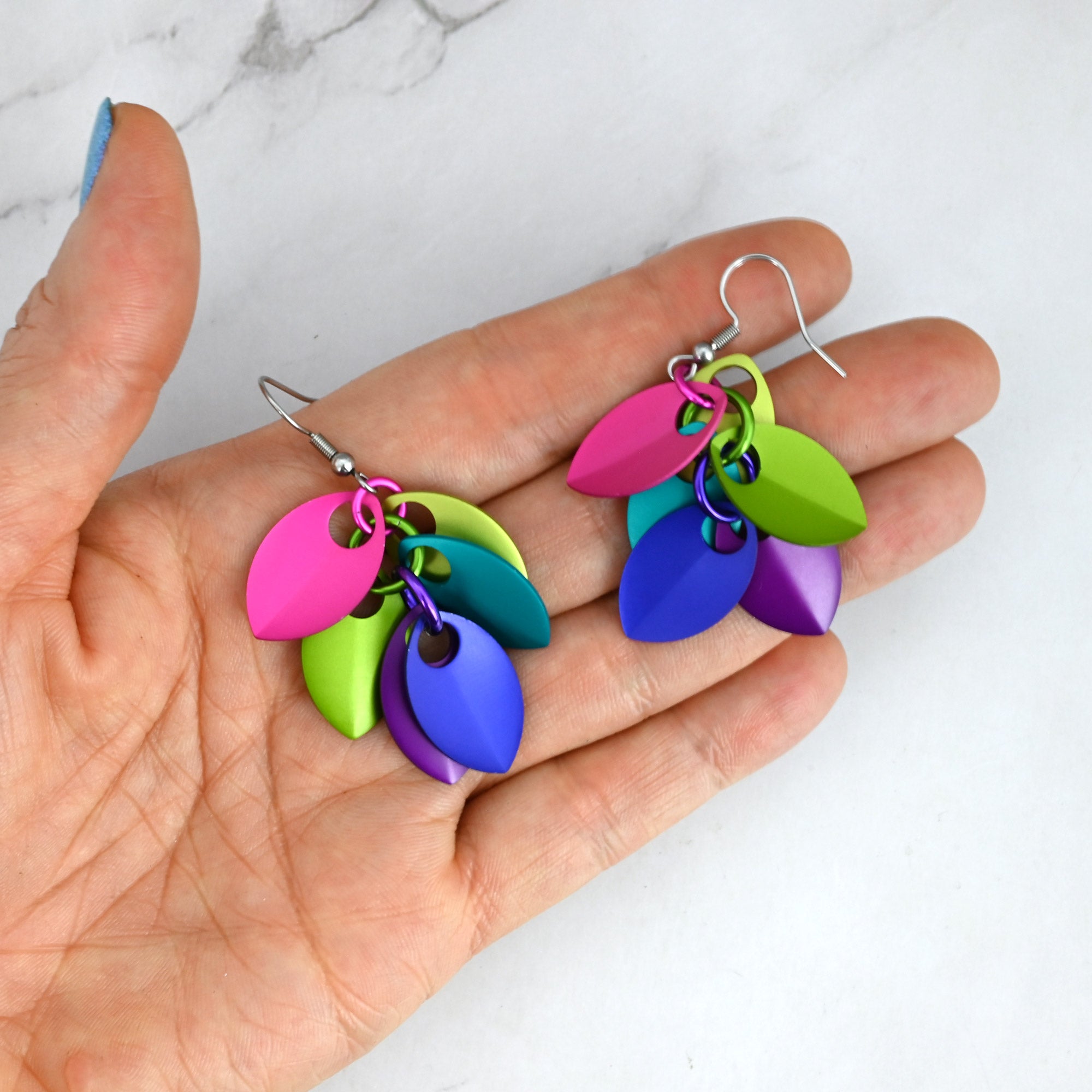 Vibrant electric rainbow scale earrings resting in a person's palm to show the vibrant, almost neon colors of the leaf-shaped components and the overall size of the earrings.