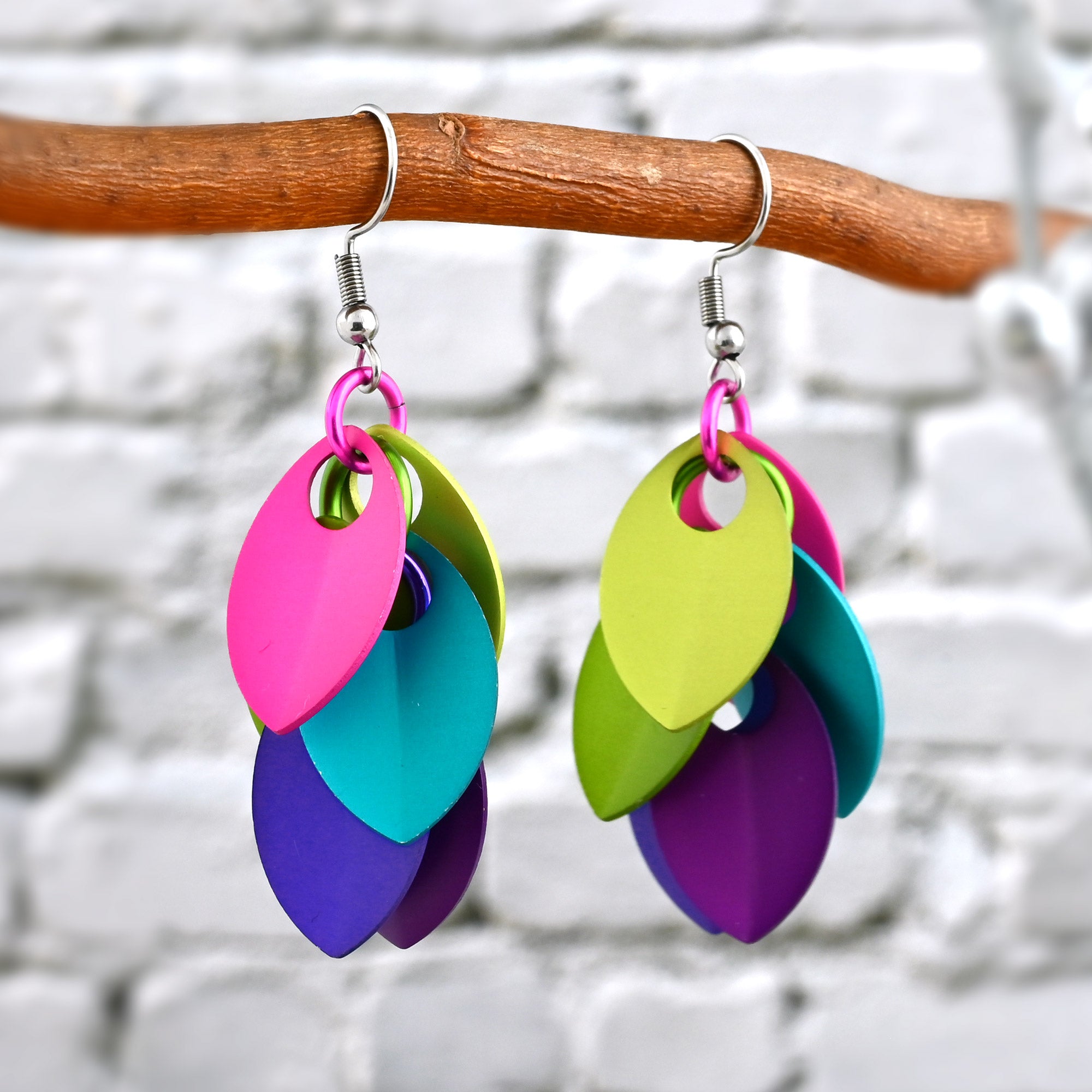 A pair of electric rainbow scalemaille earrings hanging from a branch, highlighting the bright colors against a white brick background.