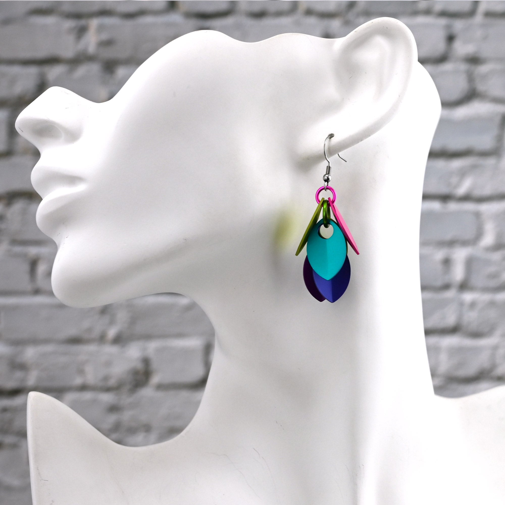 Colorful leaf-shaped earrings on a mannequin ear against a gray brick wall.