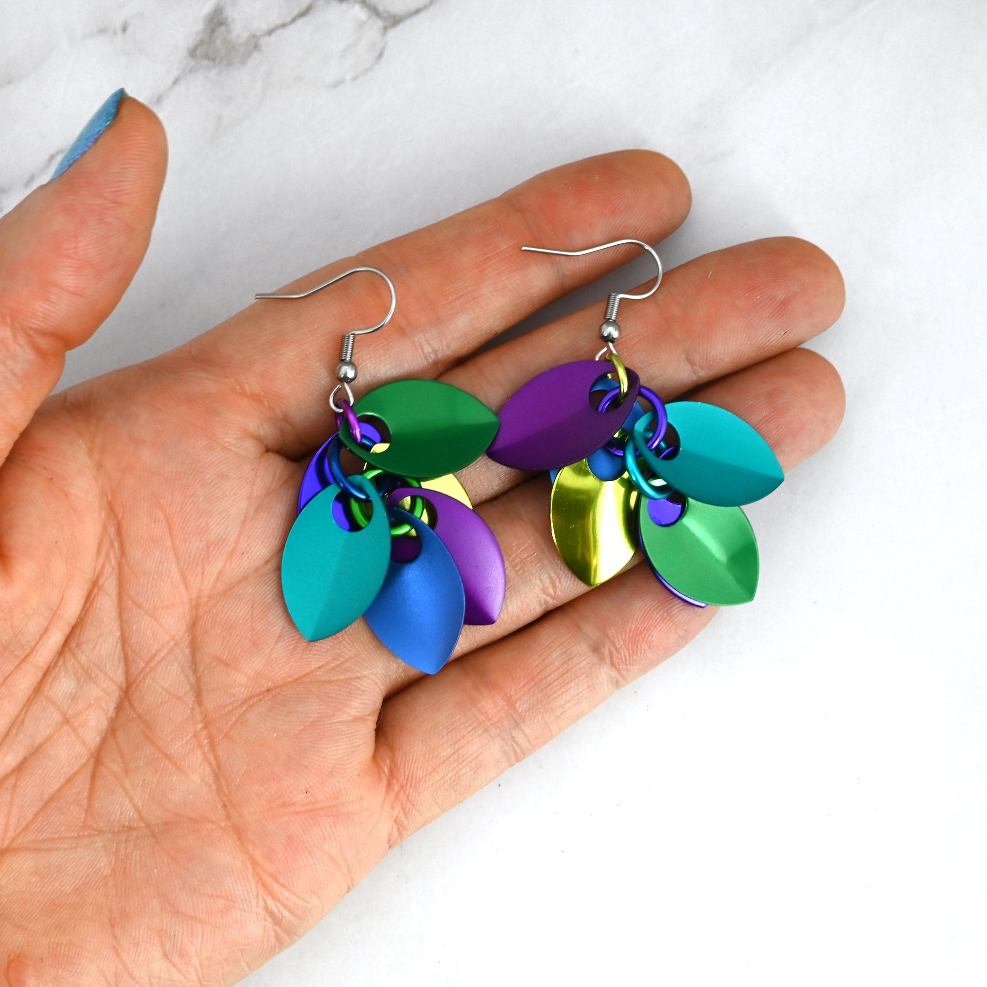 A person holding the mismatched peacock scale earrings to demonstrate their small, lightweight size.