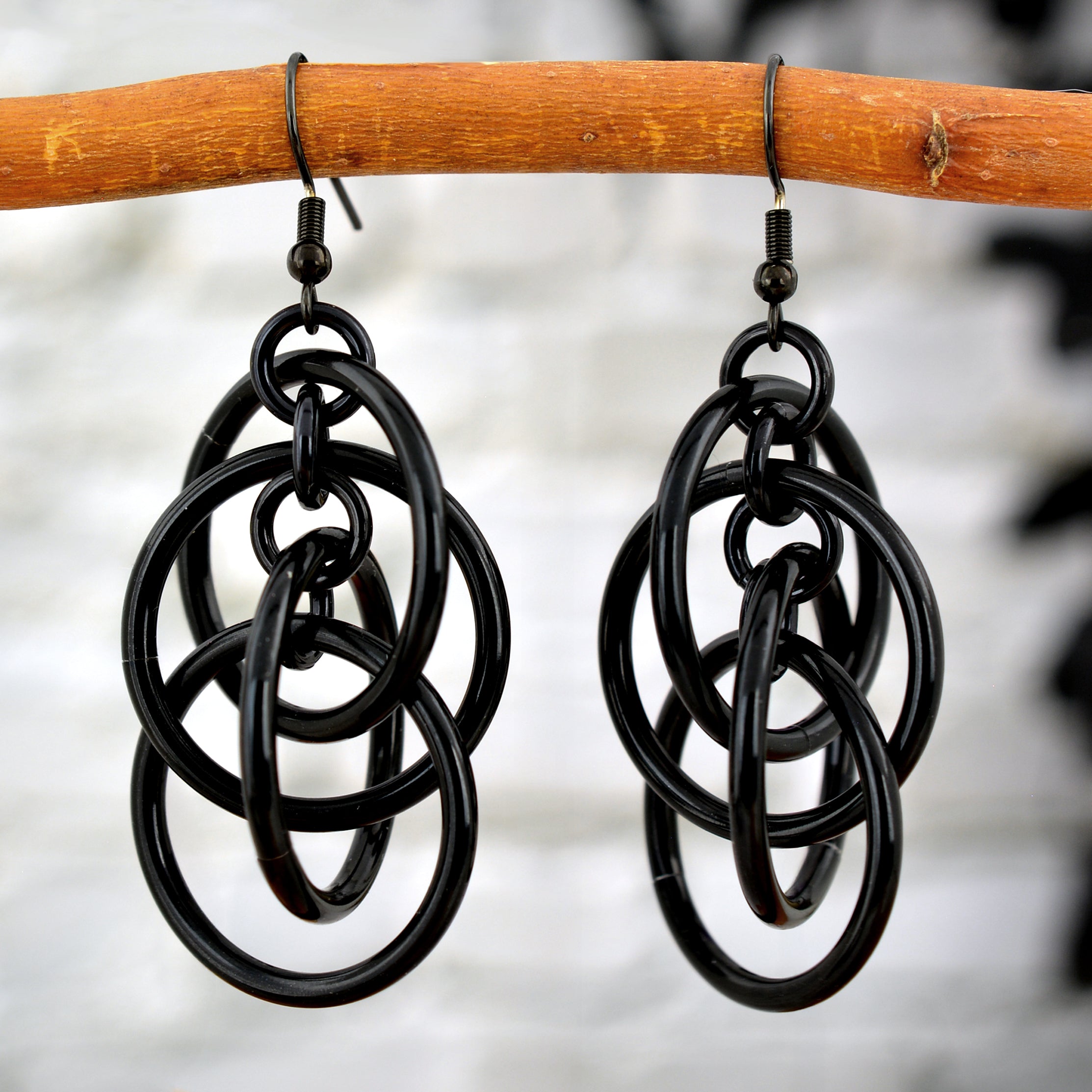Black intertwined hoop chainmaille earrings hanging from a wooden branch, highlighting the glossy finish of the rings.