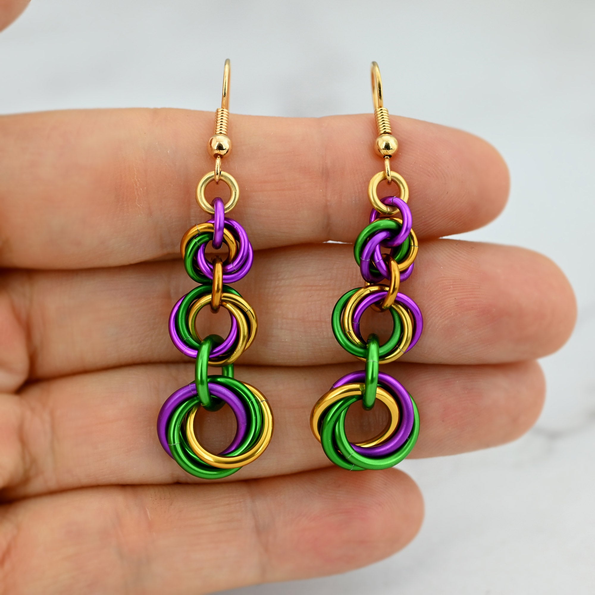 Graduated purple, gold, and green chainmaille earrings held against fingertips to show the detailed "knot" texture and size.