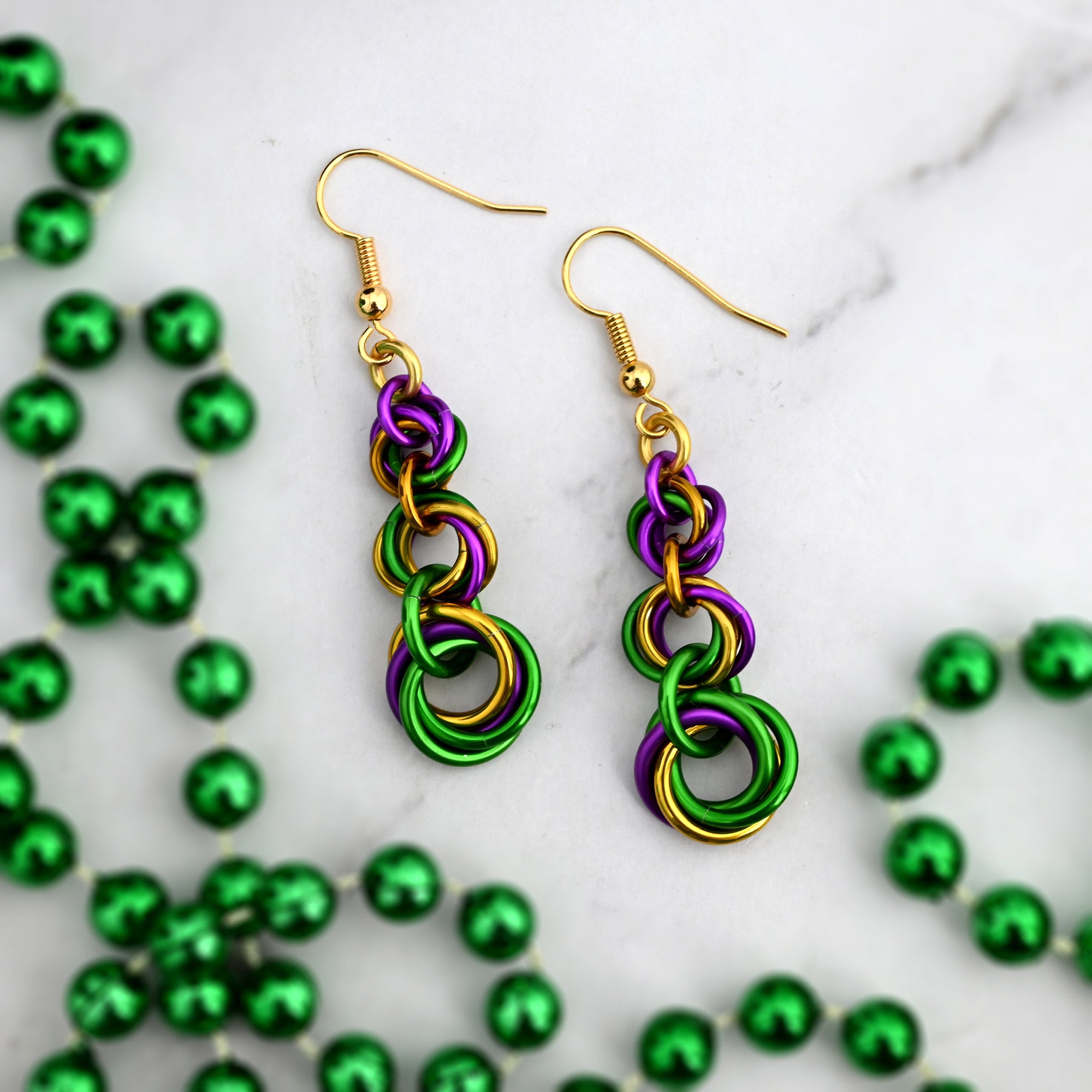 Flat lay of King Cake-inspired graduated knot earrings on white marble, accented by a string of green metallic Mardi Gras beads.