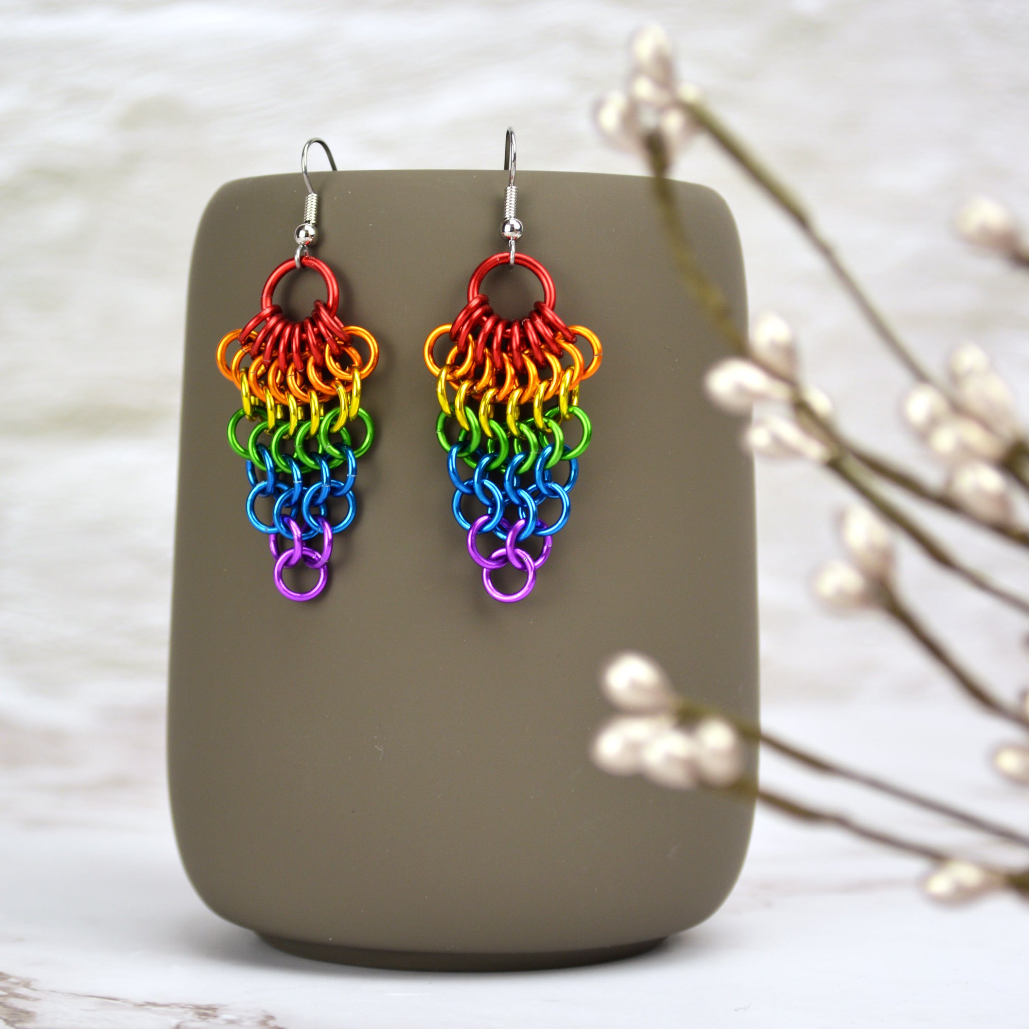 A pair of rainbow Mesh Cluster chainmaille earrings hanging from the side of a gray ceramic mug.