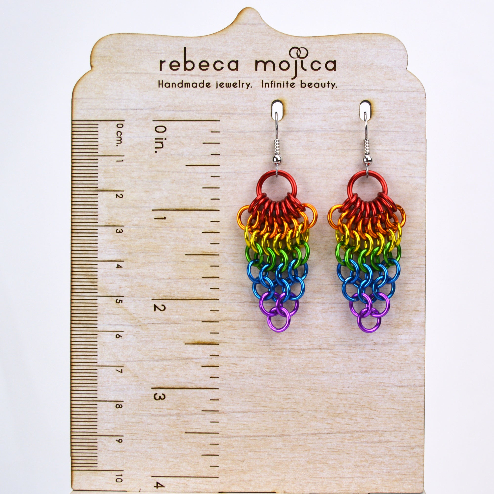 A pair of rainbow pride Mesh Cluster earrings on a wooden earring card with both an inch and a centimeter ruler, showing their size, which is just short of 2.5 inches.
