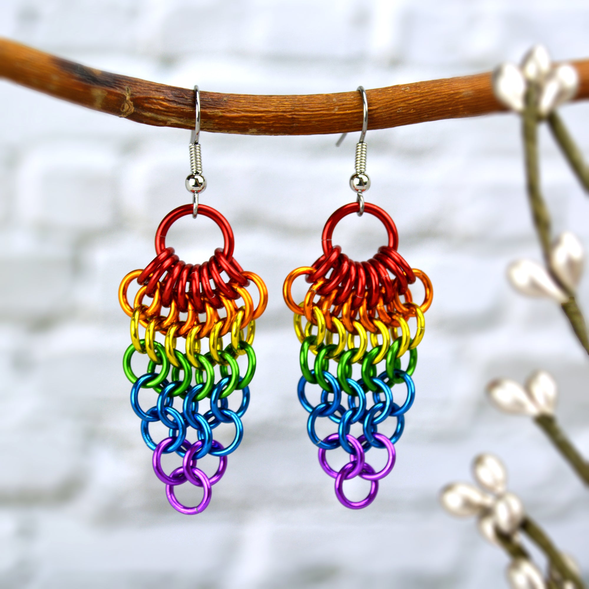 A pair of rainbow Mesh Cluster chainmaille earrings hanging from a small brown branch.