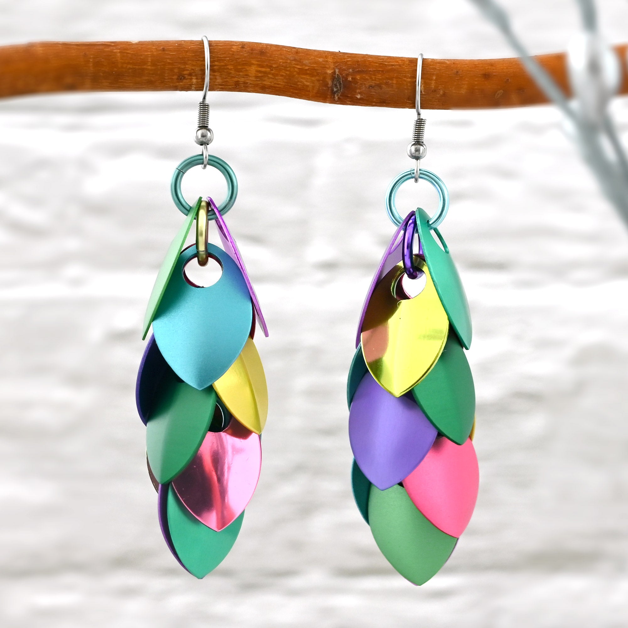 A pair of long pastel scalemaille earrings hang from a branch, highlighting the mix of matte and shiny finishes and the variety of colors. Each earring has 6 pairs of scales (leaf shaped) joined by a single chain of colorful jump rings hidden in the center