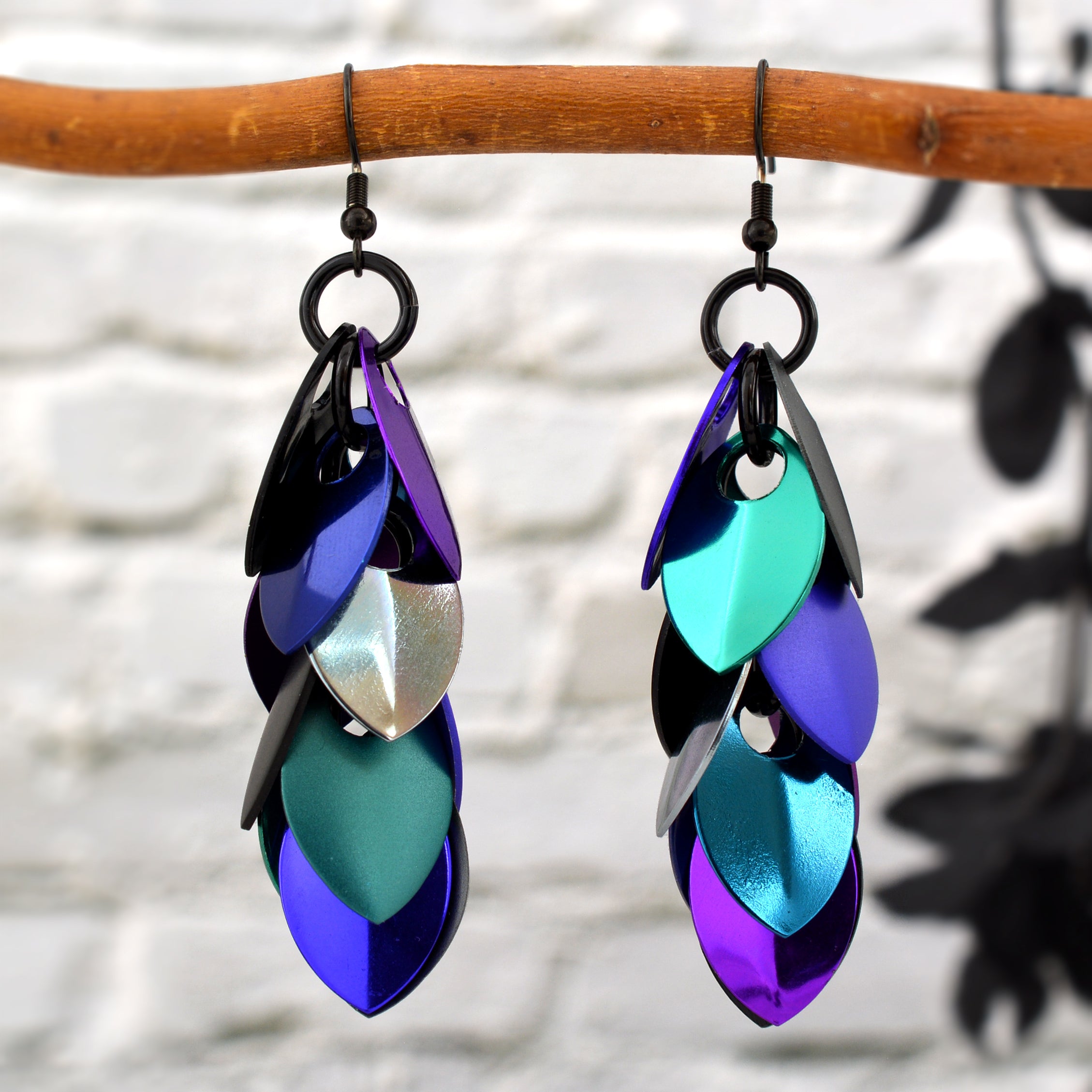A pair of Rebeca Mojica's long, colorful "Dark Mermaid" feathered chainmail earrings, with iridescent blue, purple, and teal scales, hanging from a natural wooden branch against a white brick wall.