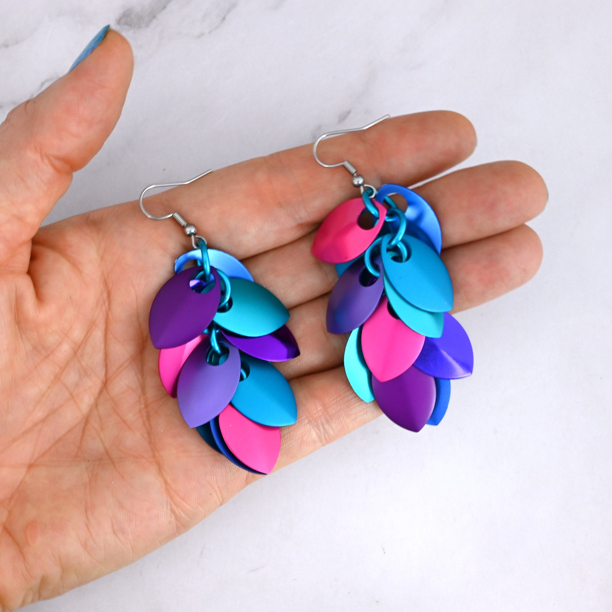 A pair of long, vibrant scalemaille earrings resting in a person’s fingertips to demonstrate their dramatic length and vivid colors, featuring shades of blue and purple plus hot pink.