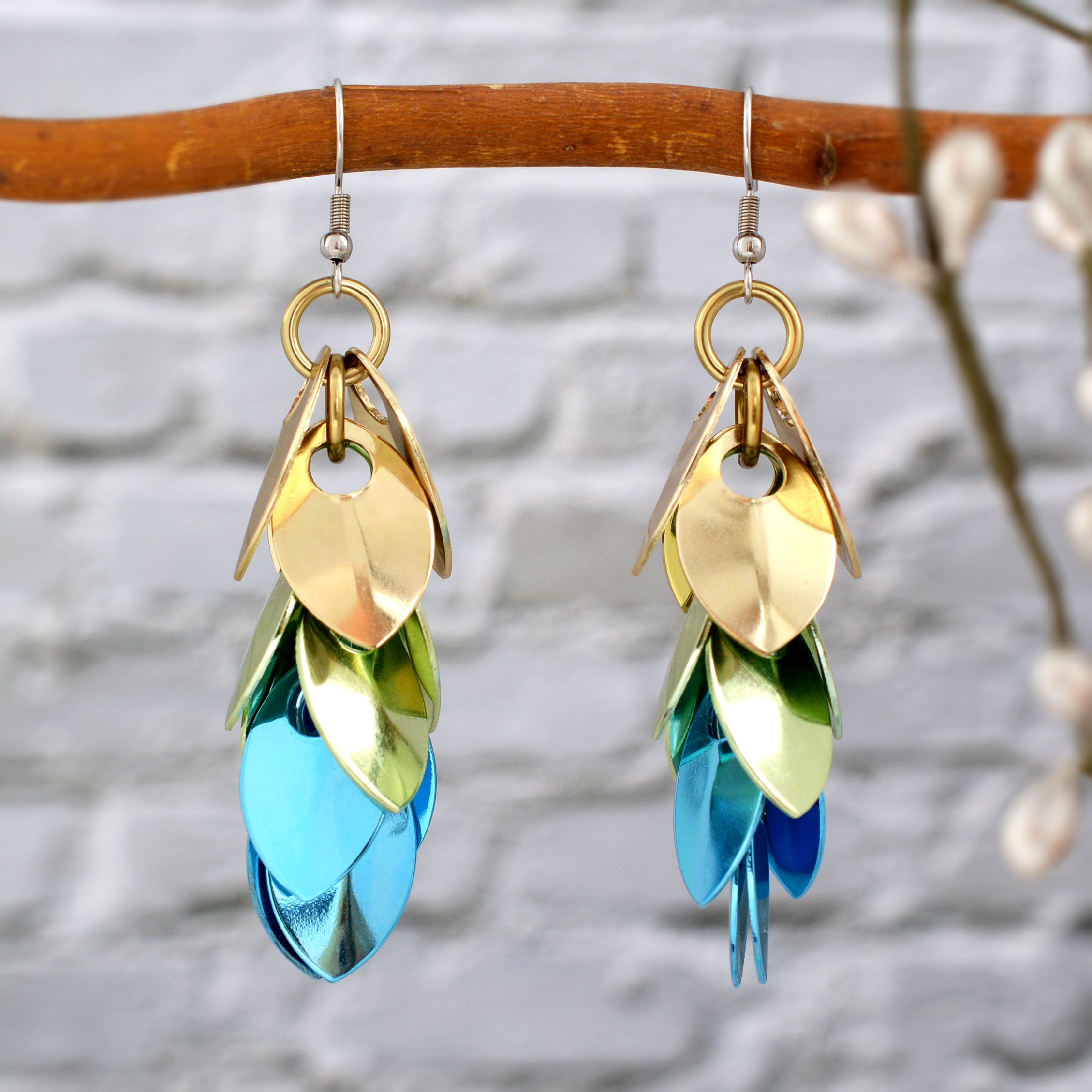 Rebeca Mojica's Cascading Leaves scalemaille earrings hang from a brown branch against a light brick wall. The shiny champagne, seafoam, and light blue scales reflect light, making the earrings stand out.