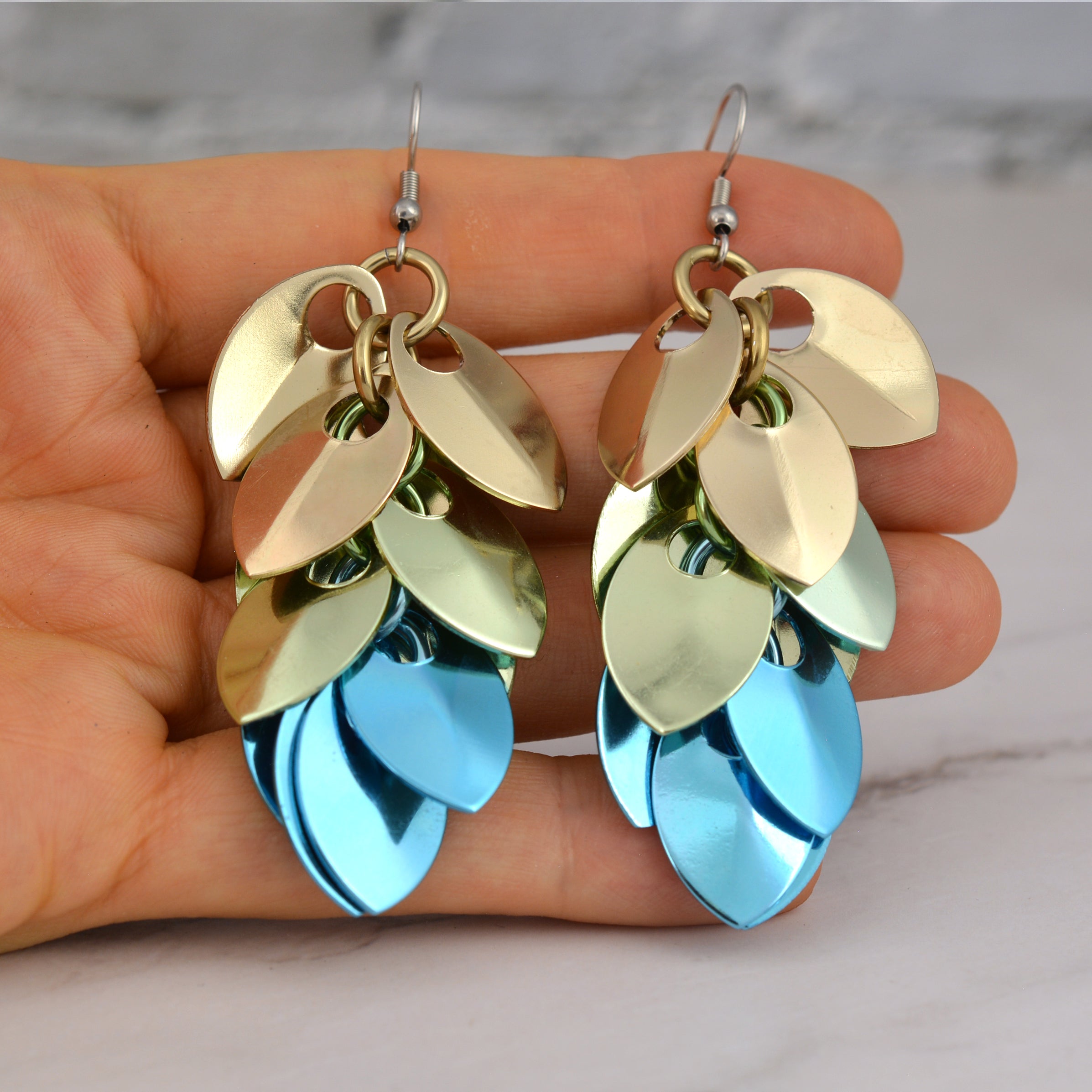A pair of scalemaille earrings resting on the palm of artisan Rebeca Mojica’s hand, showing the overall size and how the colors shimmer. A white marble surface is in the background.