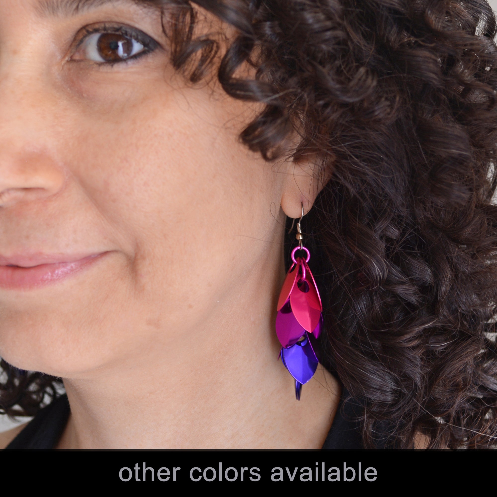 A close-up profile shot of a person with dark, curly hair wearing a long, feathered-style earring from the "Petunia" colorway. The earring is handcrafted from layered metal scales in a gradient of hot pink, violet, and purple. It hangs from a silver-tone ear wire and rests against the person's neck. The bottom of the image features a black banner with the text "other colors available" in a white sans-serif font.