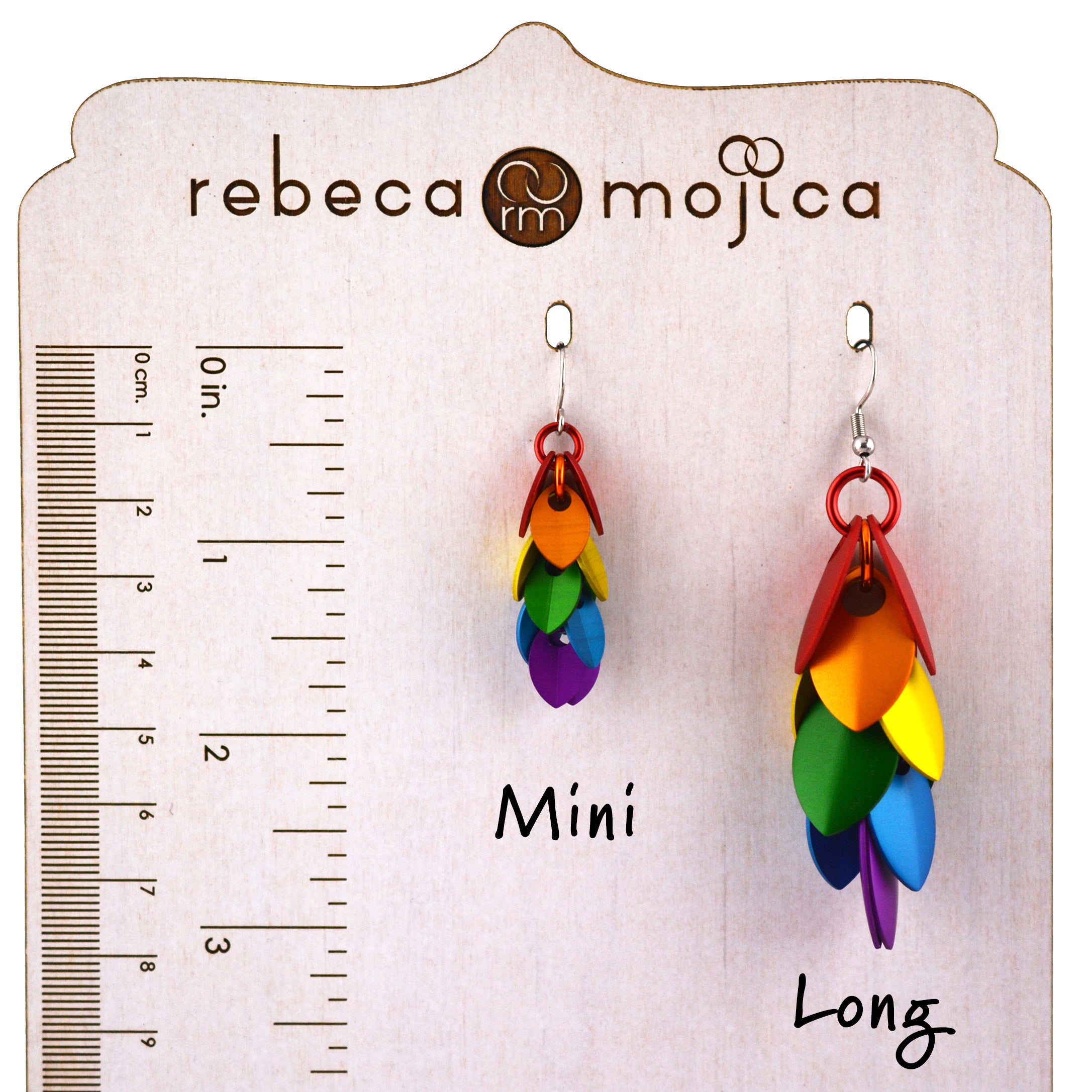 Two pairs of Rebeca Mojica Jewelry's rainbow pride earrings displayed side-by-side on a branded measuring board. On the left is the smaller "Mini" size, and on the right is the longer "Long" cascading leaves style, both featuring colorful aluminum leaves.