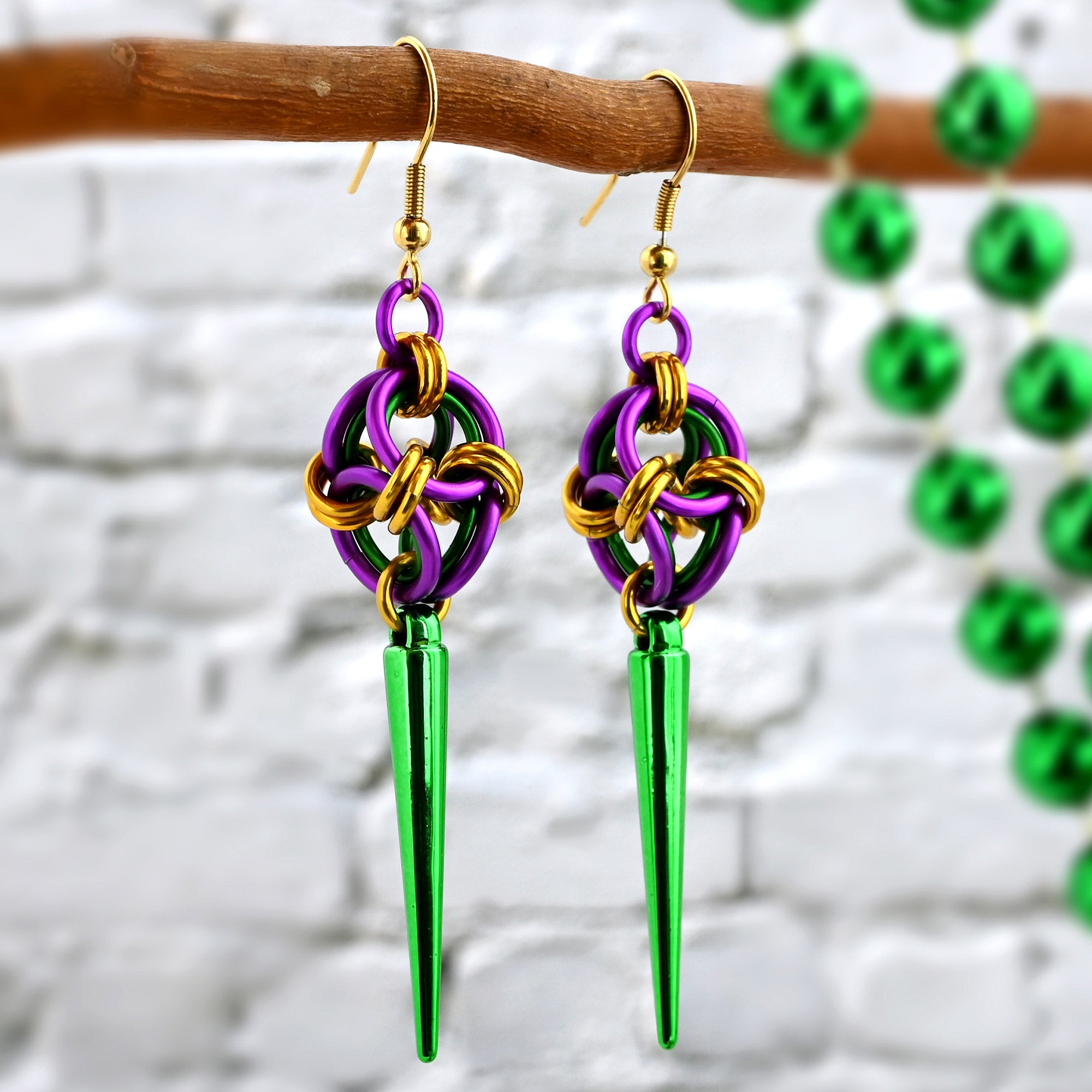 Close-up of chainmaille earrings in Mardi Gras colors (purple, green, and gold) finished with a long green spike. The earrings hang from a natural wood branch.