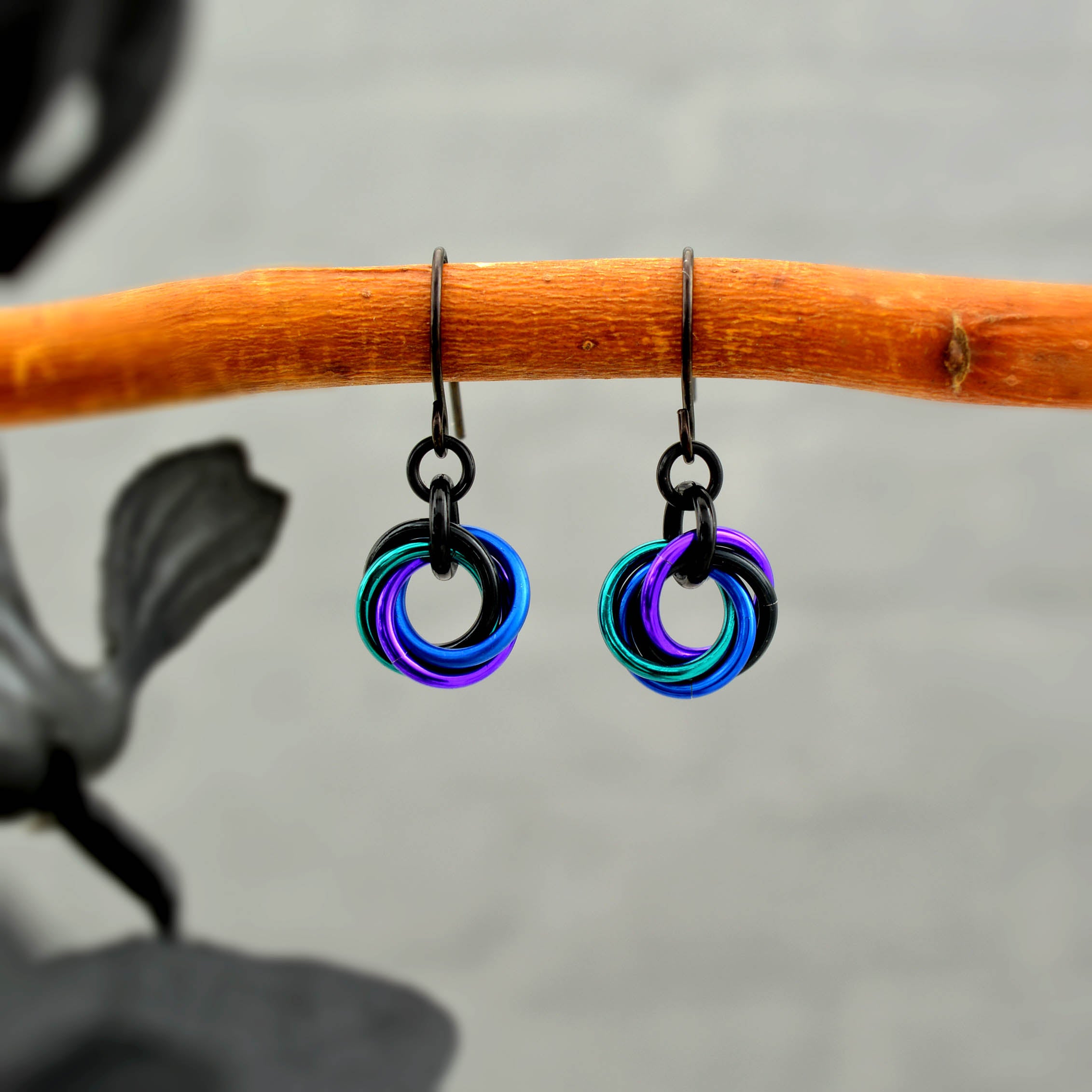 Pair of Mini Knot chainmaile earrings in a vibrant swirl of teal, blue, purple, and black rings, hanging from a natural wooden branch.
