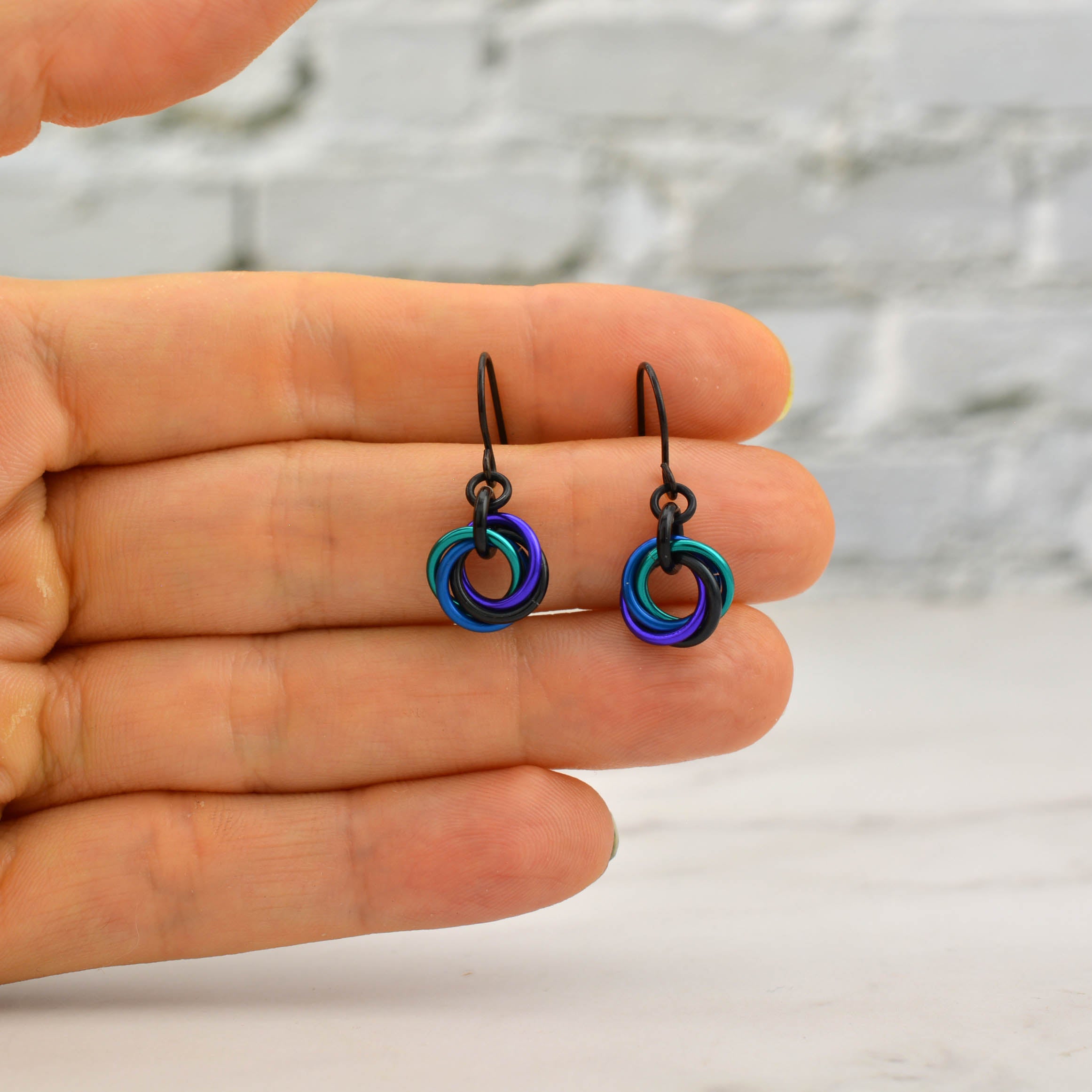 Artisan Rebeca Mojica holding a pair of tiny Mini Knot chainmaile earrings in her fingertips for size scale. Features teal, blue, purple, and black rings.
