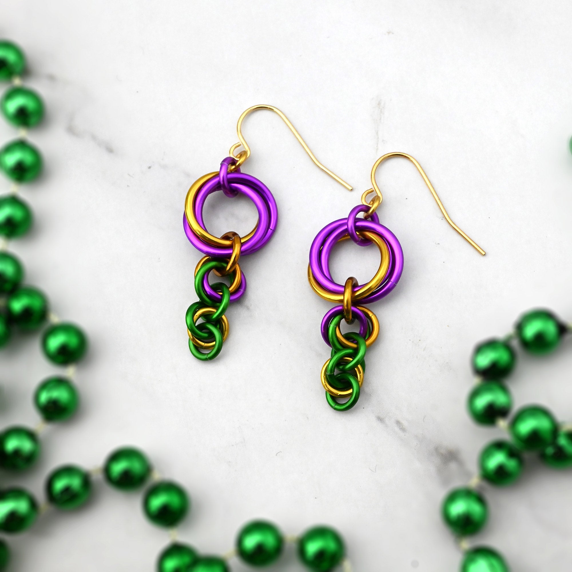 Flat lay of King Cake-inspired chainmaille knot earrings in purple, gold, and green on white marble with festive green beads.
