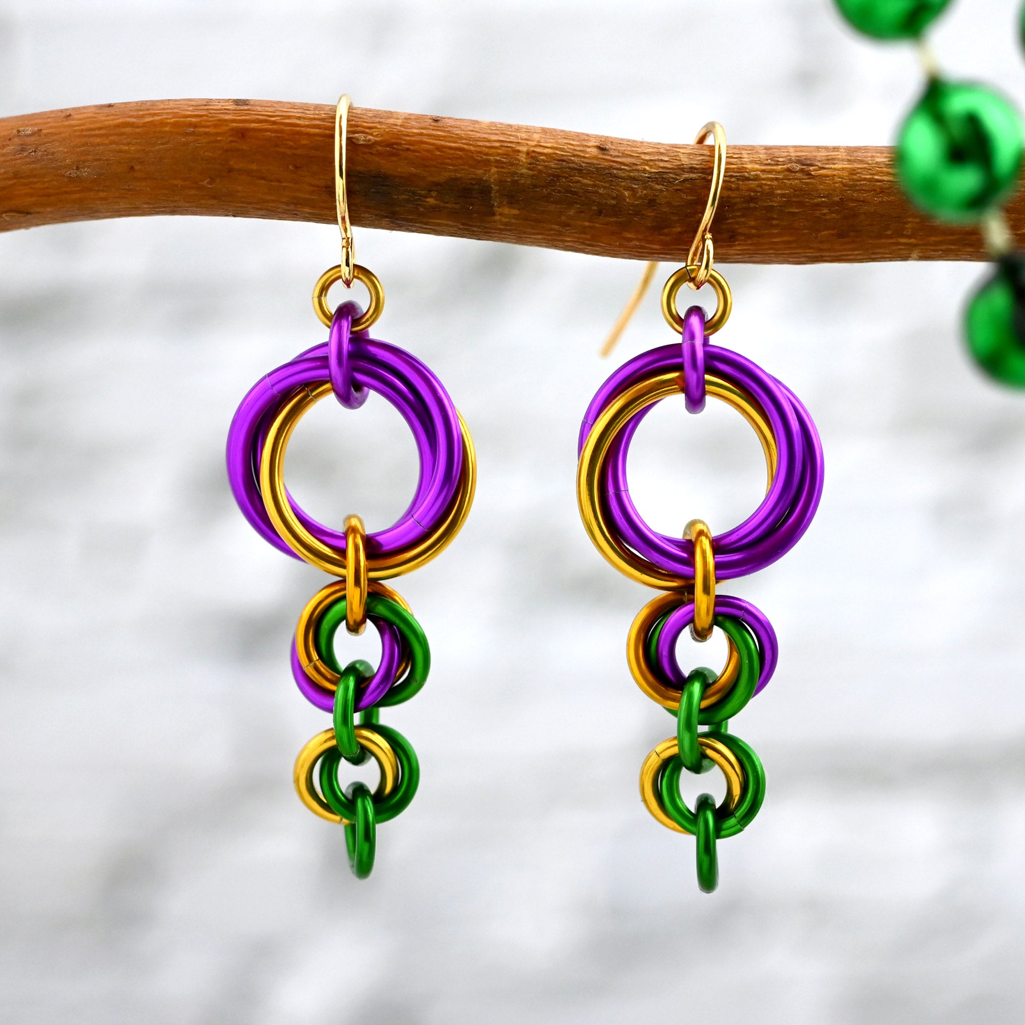 Close-up of swirly violet, gold, and green chainmaille dangles hanging from a rustic wooden branch. Top swirl has 2 large violet links and a gold link, next swirl has one of each gold, green and violet links in a smaller size, and the bottom swirl has a gold and a green link in an even smaller size. A single green link dangles from the bottom.