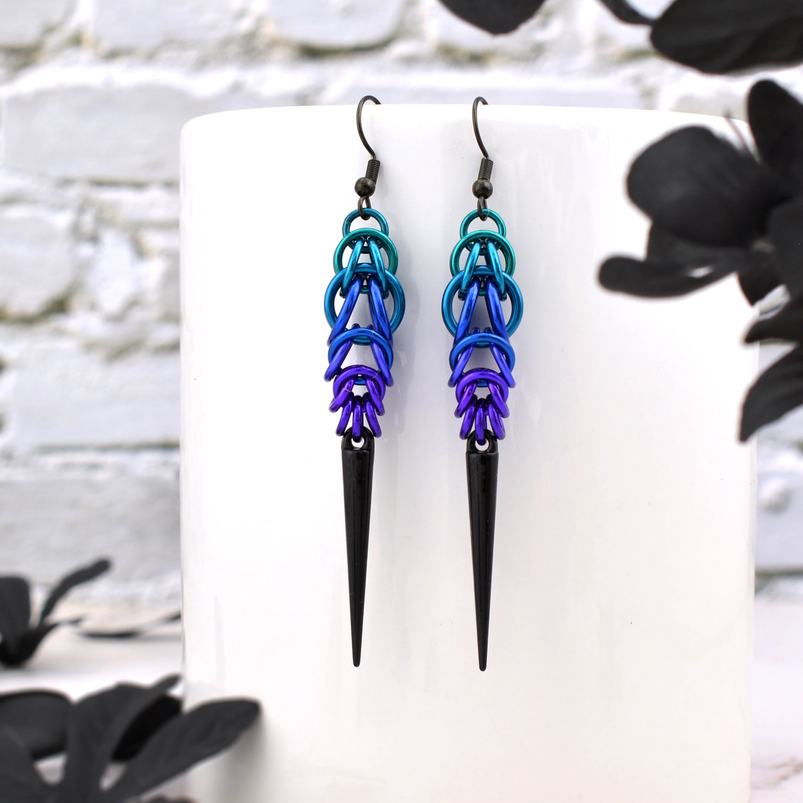 A pair of Goth-style spike earrings in teal, blue, purple, and black chainmaile, hanging on a white mug against a white brick background.