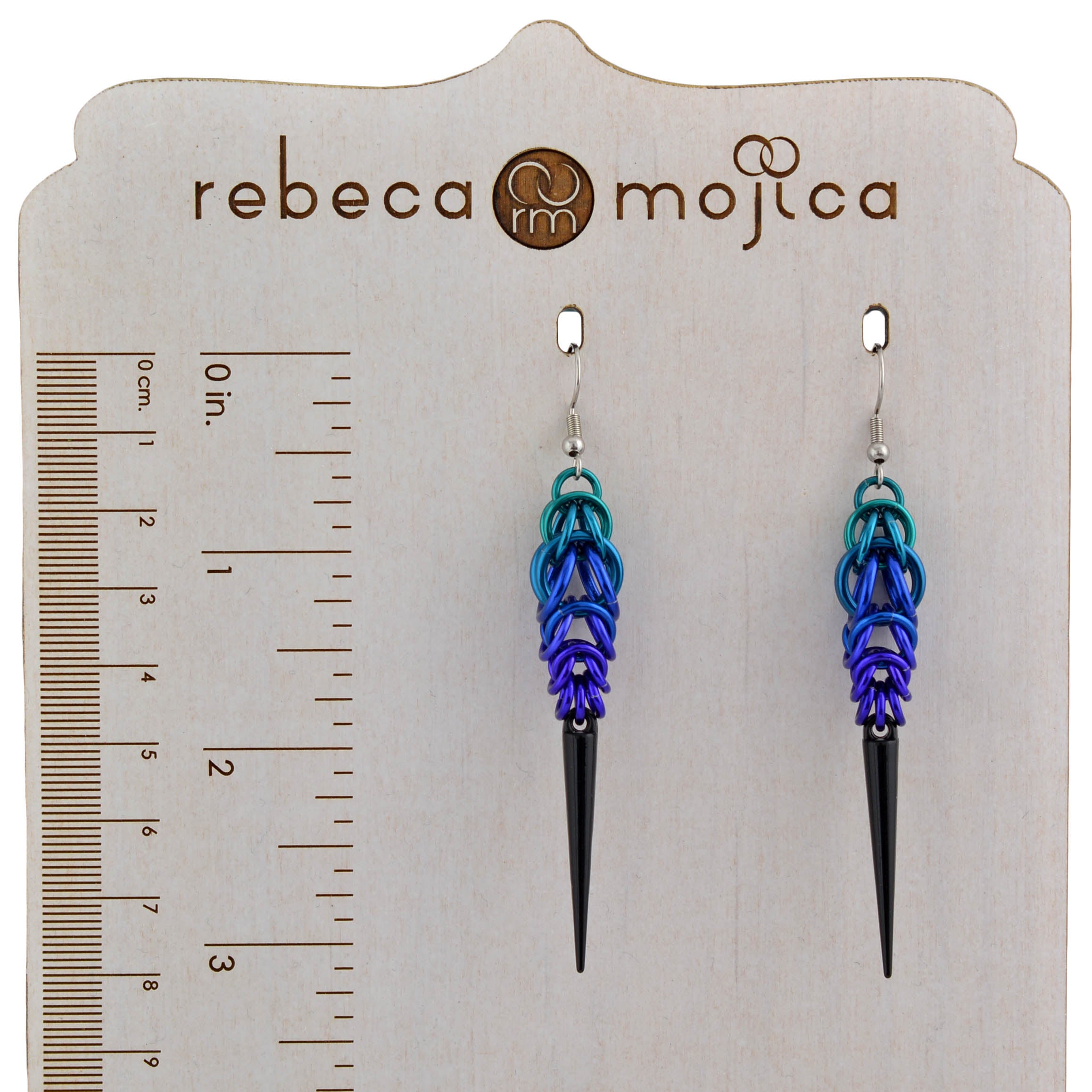 Size comparison for the Dark Mermaid Spike Earrings (approximate 3-inch length) on the Rebeca Mojica branded wooden display with ruler.