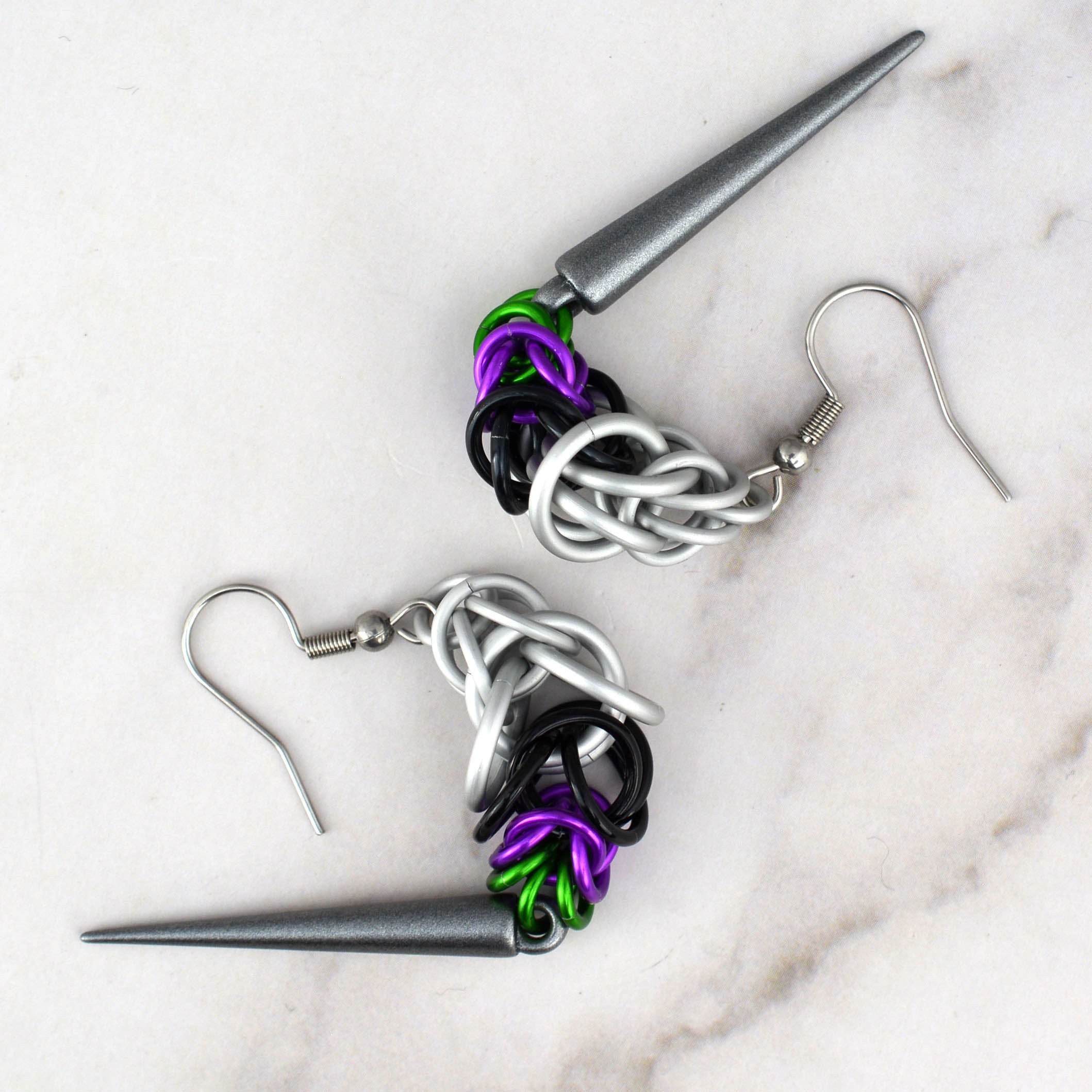 A pair of Double Demi Pride chainmaille earrings featuring white, black, violet, and green rings, with a dark grey spike. The earrings are curved away from each other on a white marble surface, highlighting the flexibility of the chainmaille weave.