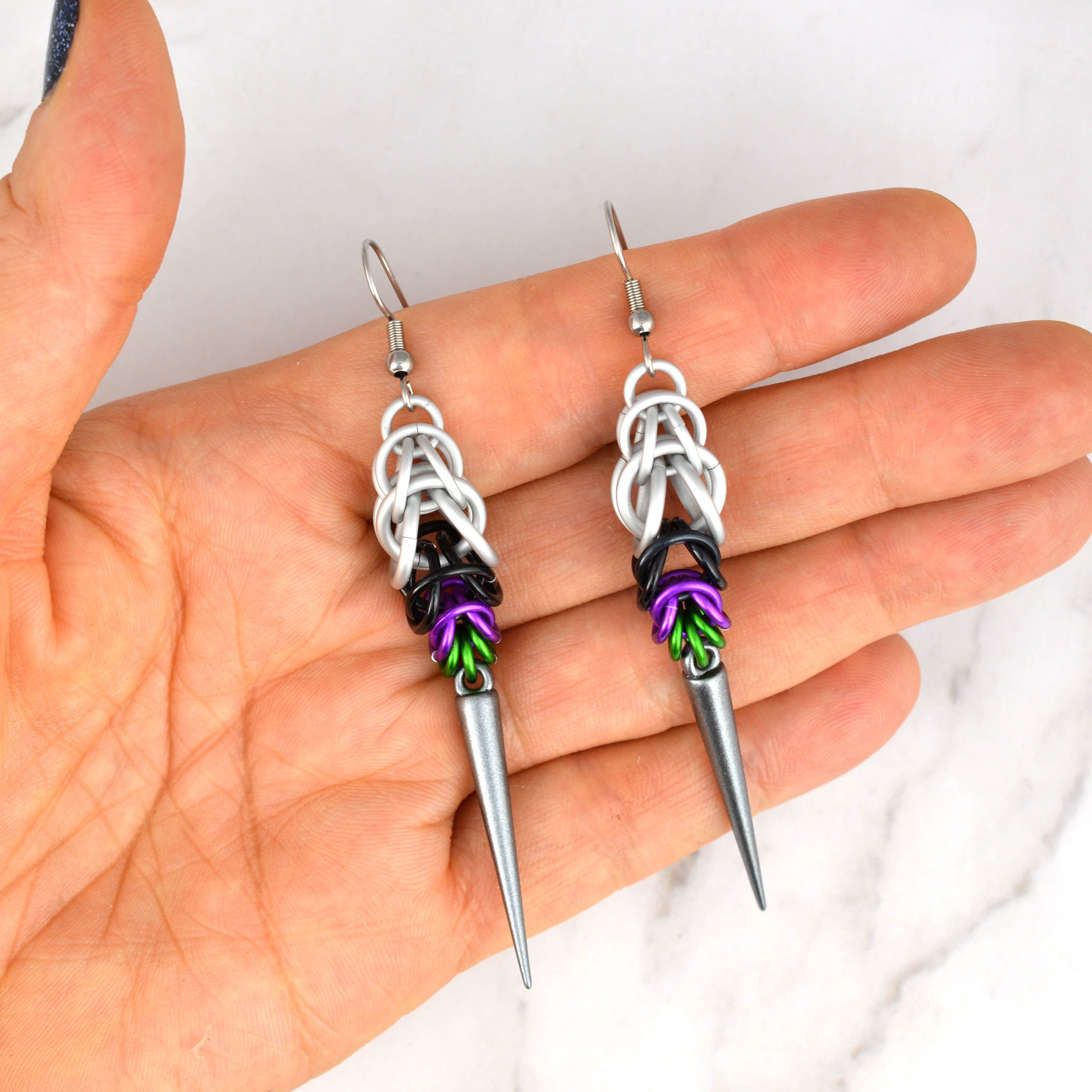 A pair of Double Demi Pride chainmaille earrings, handmade by artisan Rebeca Mojica, being held in her open palm. The chainmaille features white, black, violet, and green aluminum rings, with a dark grey acrylic spike.