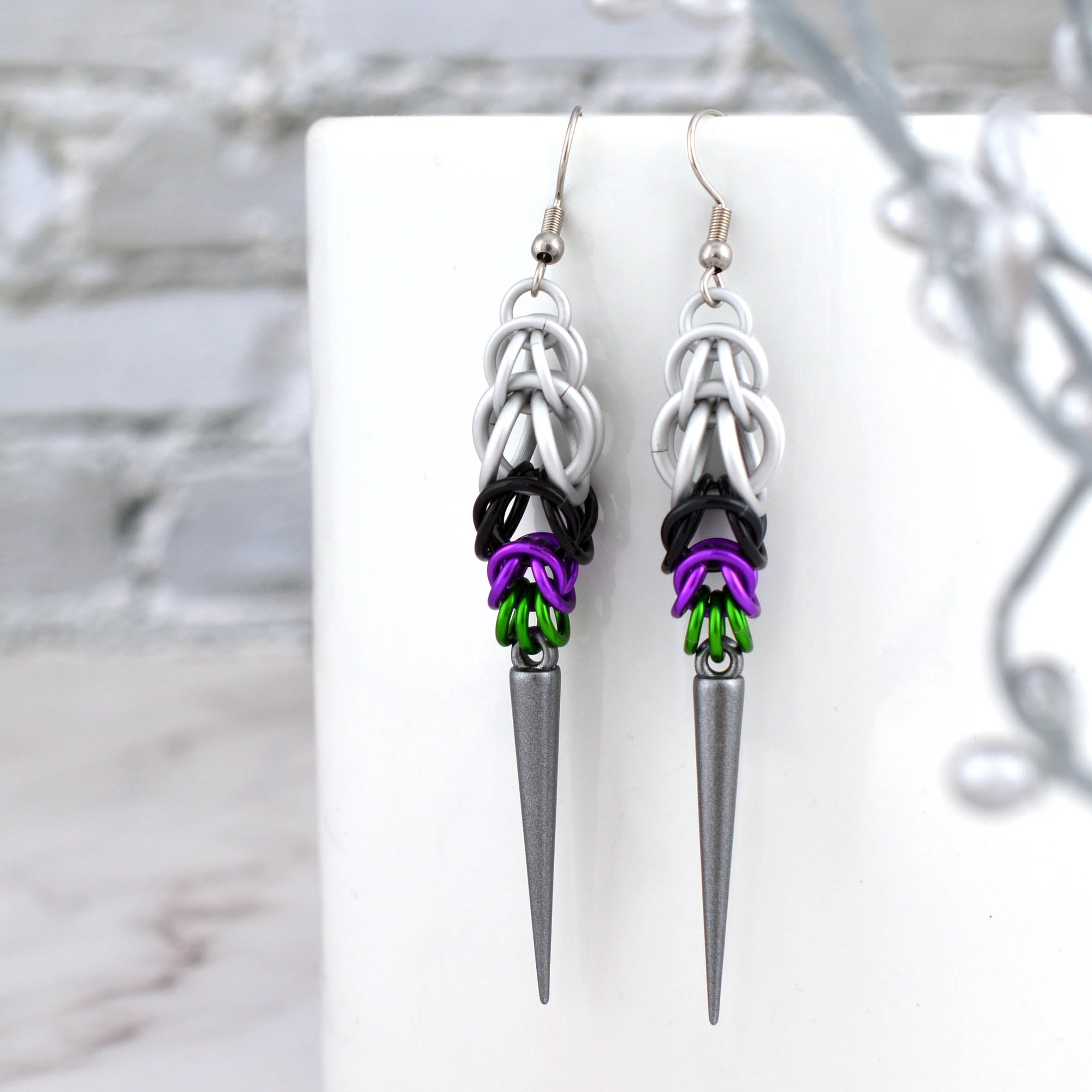 A pair of Double Demi Pride spike earrings hanging from a white ceramic mug. The chainmaille shows the pattern of white, black, violet, and green aluminum rings above a dark grey spike.