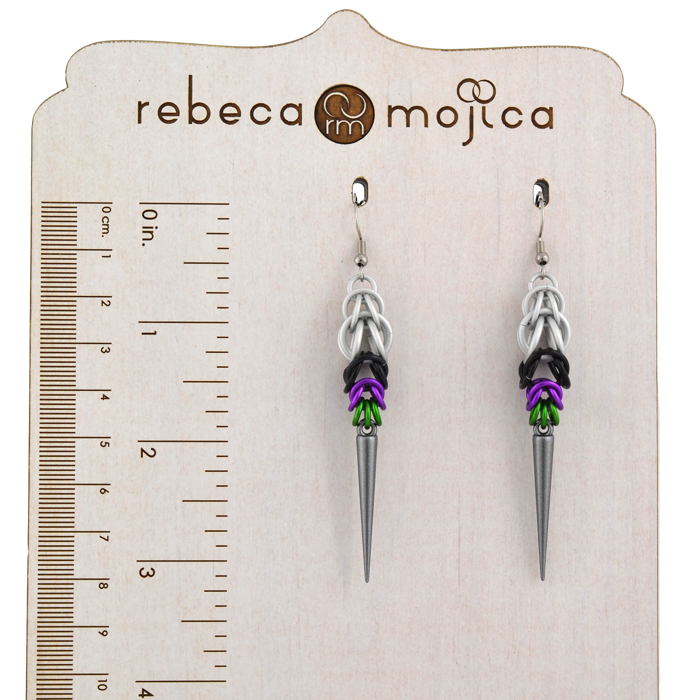 A pair of Double Demi Pride spike earrings, handmade by Rebeca Mojica, hanging from a branded wooden earring display card with a ruler graphic to show the size in inches and centimeters.