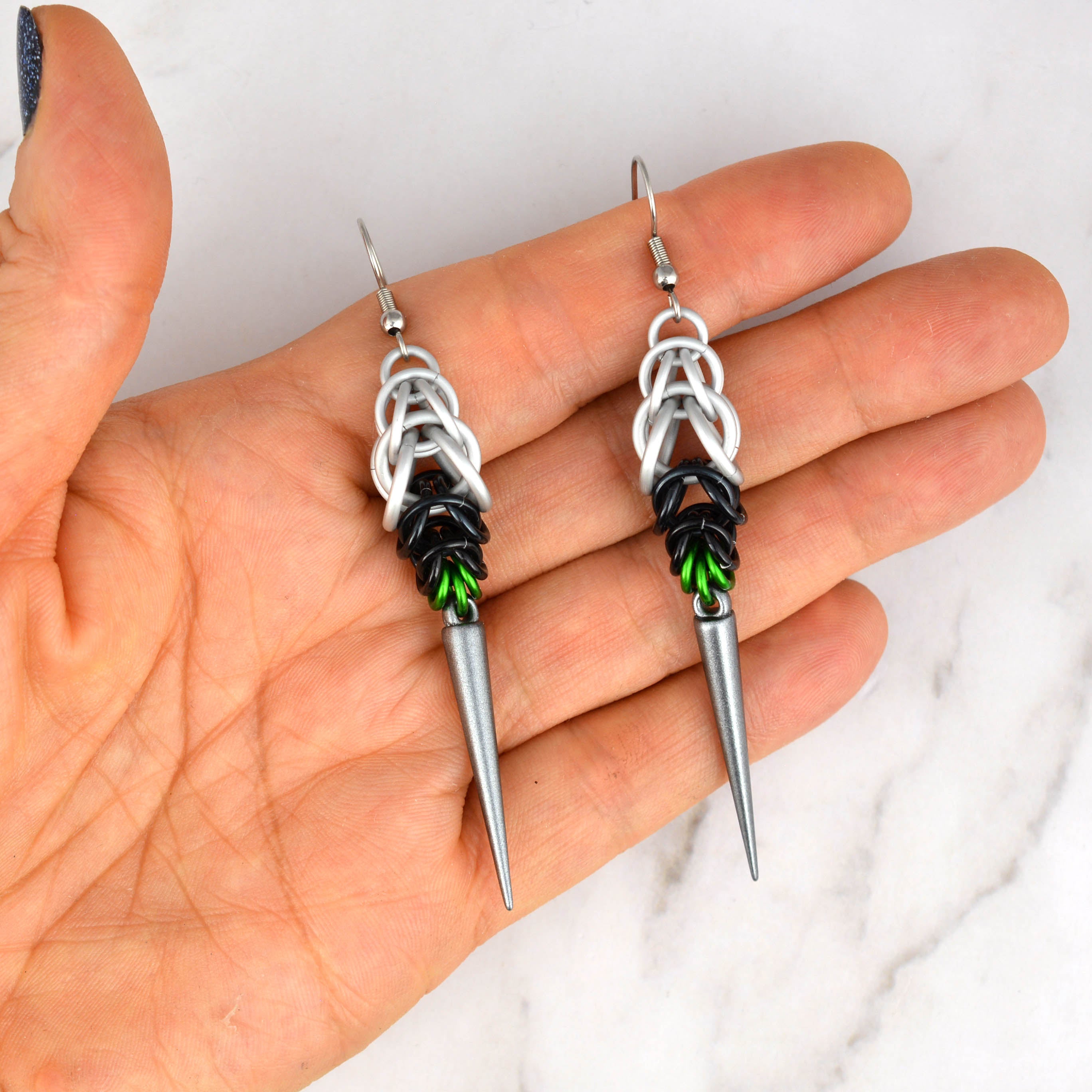 A pair of Demiromantic Pride chainmaille earrings, handmade by artisan Rebeca Mojica, being held in her open palm. The chainmaille features white, black, and green aluminum rings, with a dark grey acrylic spike.
