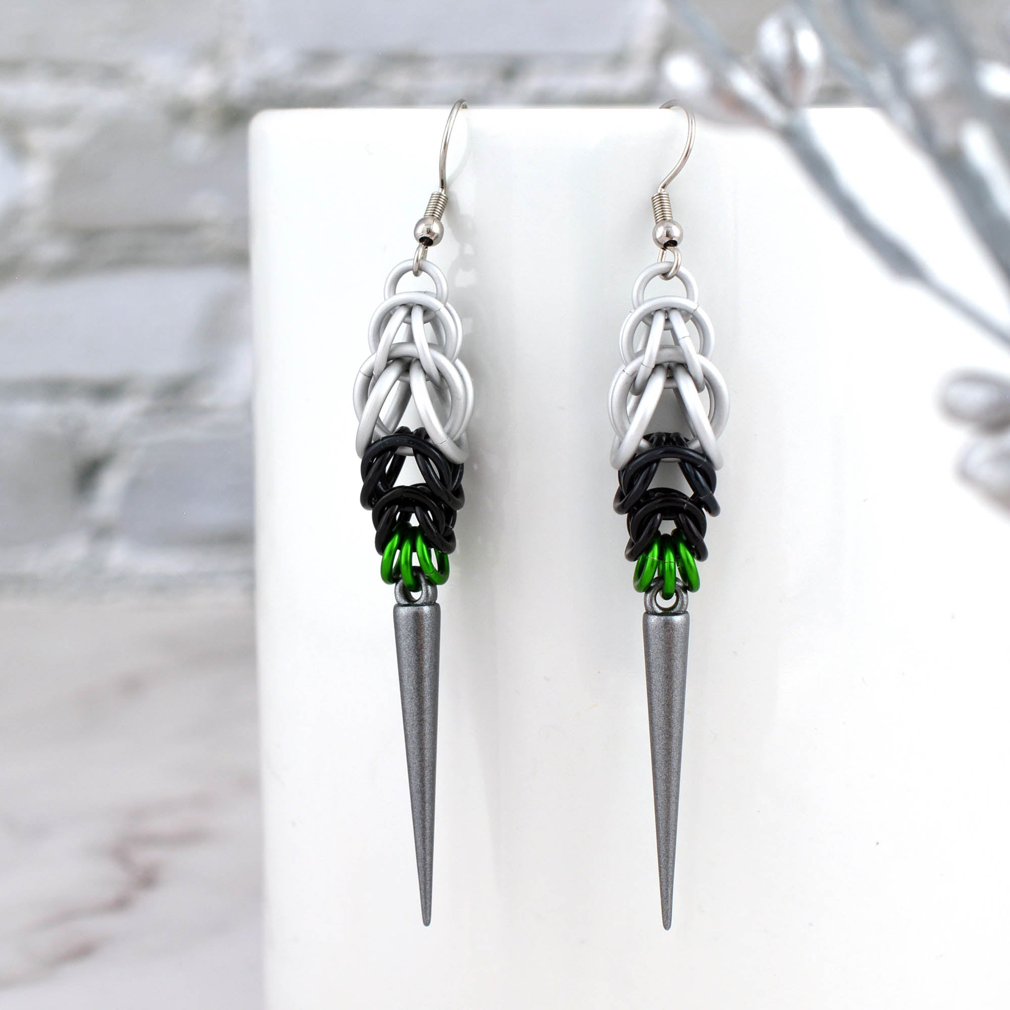 A pair of Demiromantic Pride spike earrings hanging from a white mug. The chainmaille shows the pattern of white, black, and green aluminum rings above a dark grey spike.