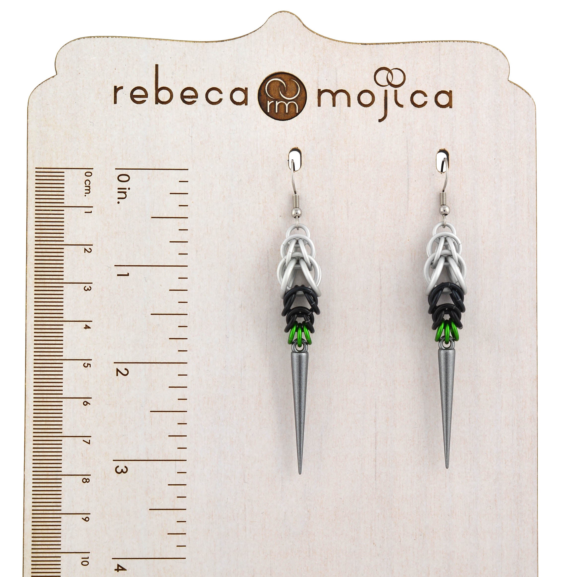 A pair of Demiromantic Pride spike earrings, handmade by Rebeca Mojica, hanging from a branded wooden earring display card with a ruler graphic to show the size in inches and centimeters.