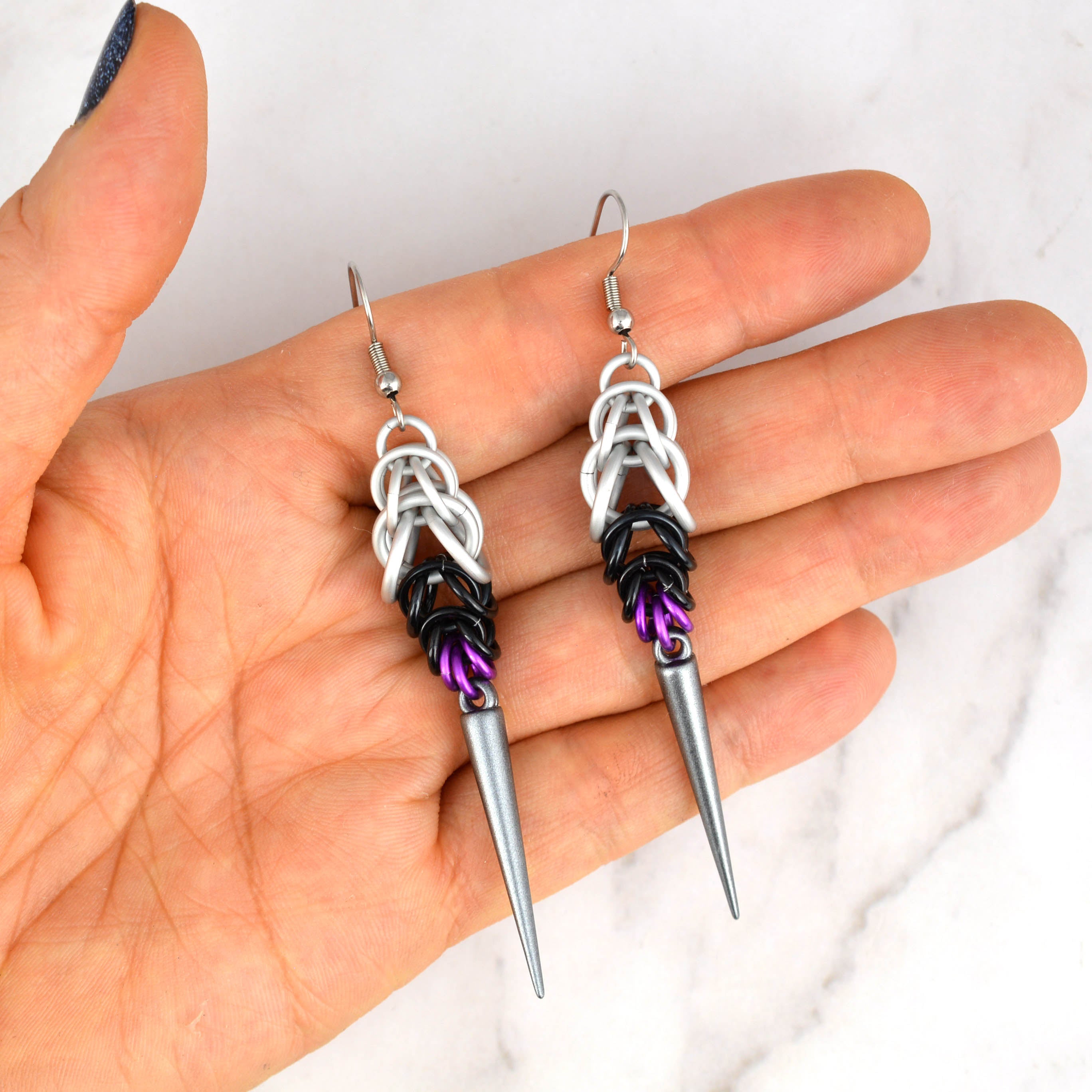 A pair of Demisexual Pride spike earrings, handmade by artisan Rebeca Mojica, being held in her open palm against a white marble background. This image shows the scale and size of the earrings relative to a person's hand.