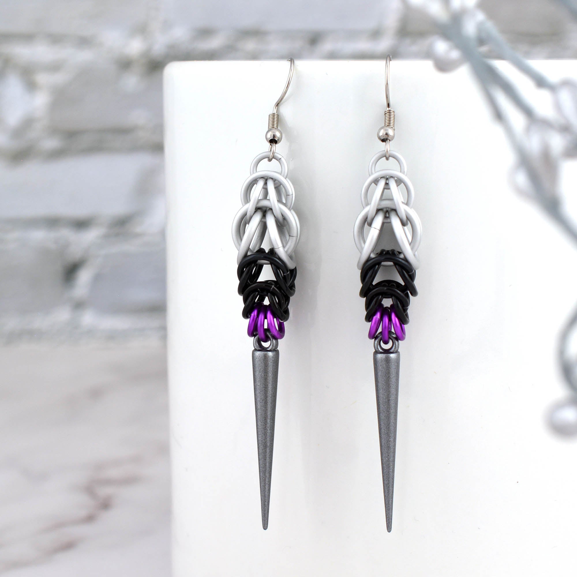 A close-up, vertical view of the pair of Demisexual Pride spike earrings hanging from the edge of a white ceramic mug. The chainmaille is clearly visible, showing the pattern of white, black and purple aluminum rings, with a dark grey spike at the base. A blurred white brick wall is in the background.