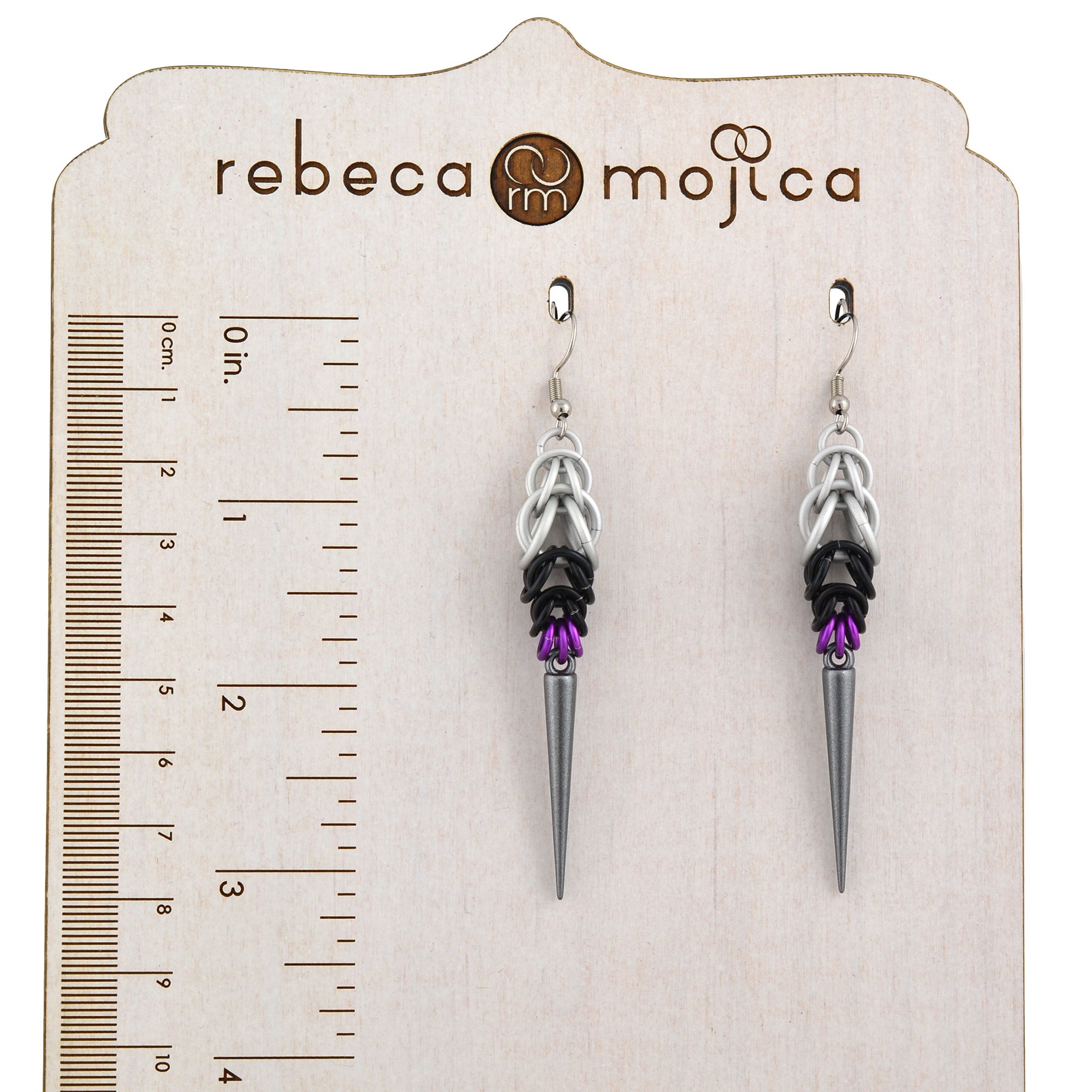 A pair of Demisexual Pride spike earrings, handmade by Rebeca Mojica, hanging from a branded wooden earring display card that features a ruler graphic on the left side, showing both inches and centimeters. This image helps customers visualize the exact size and length of the earrings, approximately 3.25 inches