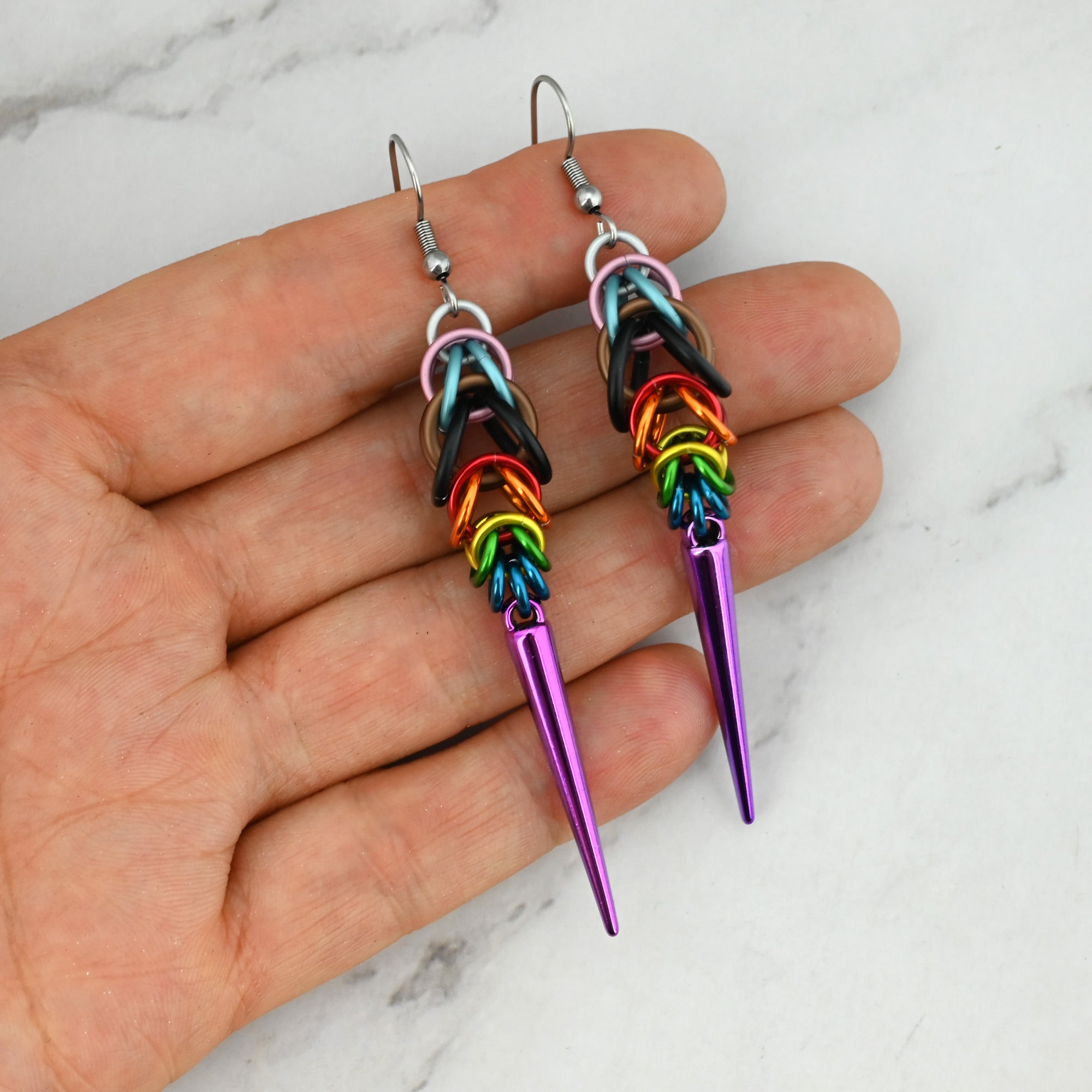 A pair of Progress Pride spike earrings held across a person's fingertips to show scale. The earrings feature colorful aluminum rings in a tapered chainmaille pattern ending in long, shiny purple spikes.