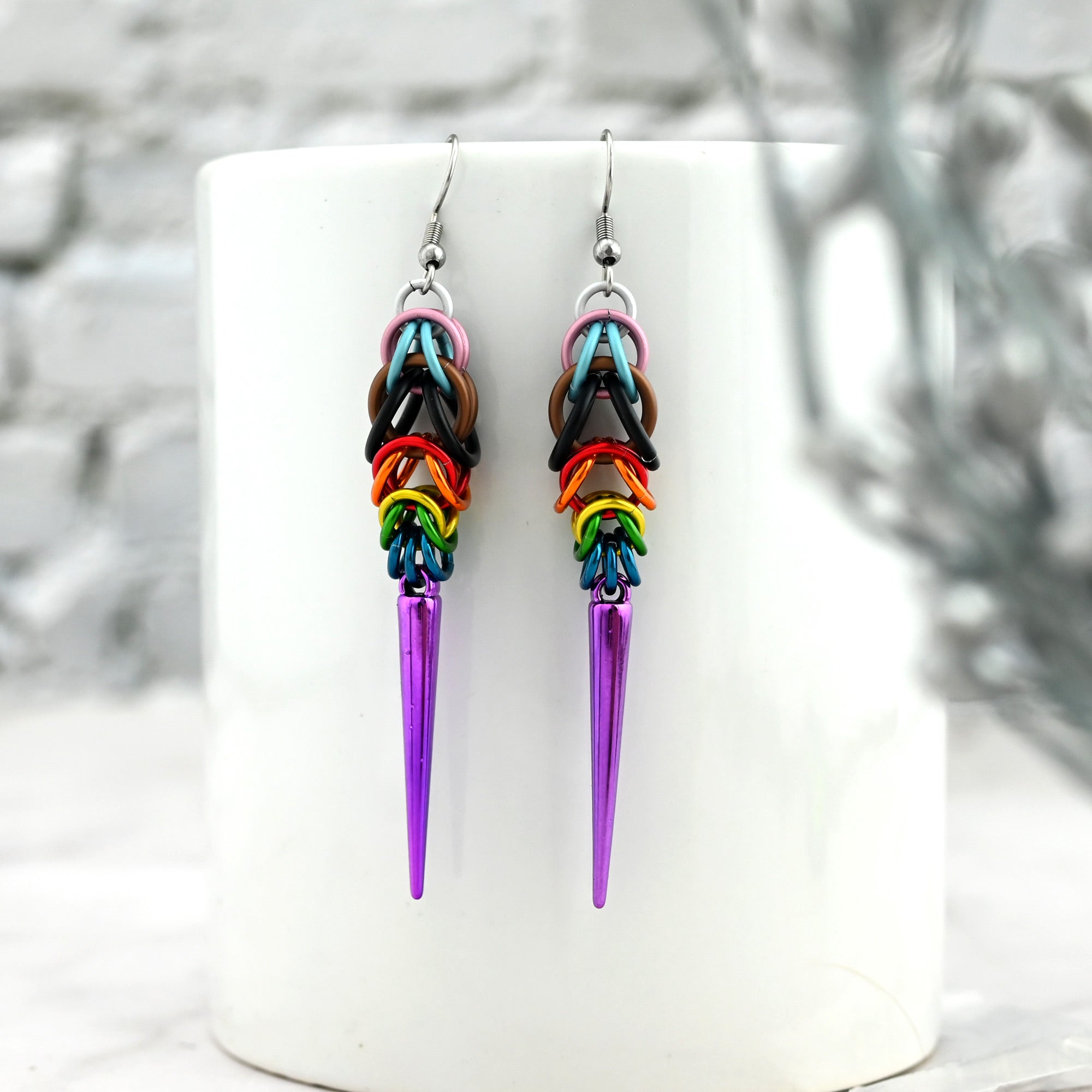 Progress Pride spike earrings draped over the rim of a clean white ceramic mug. The white background and minimal styling emphasize the brilliant colors of the chainmaille weave and the sharp, polished look of the purple spikes.