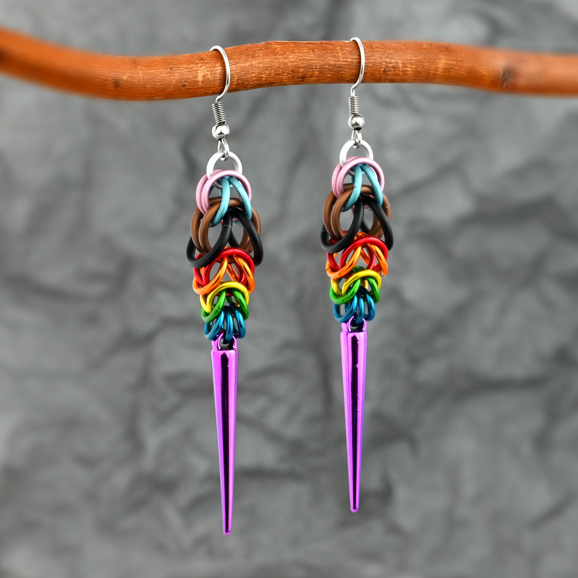 Two Progress Pride spike earrings hanging from a natural wood branch against a textured gray background. The metallic colors of the aluminum rings and purple spikes pop against the neutral tones.