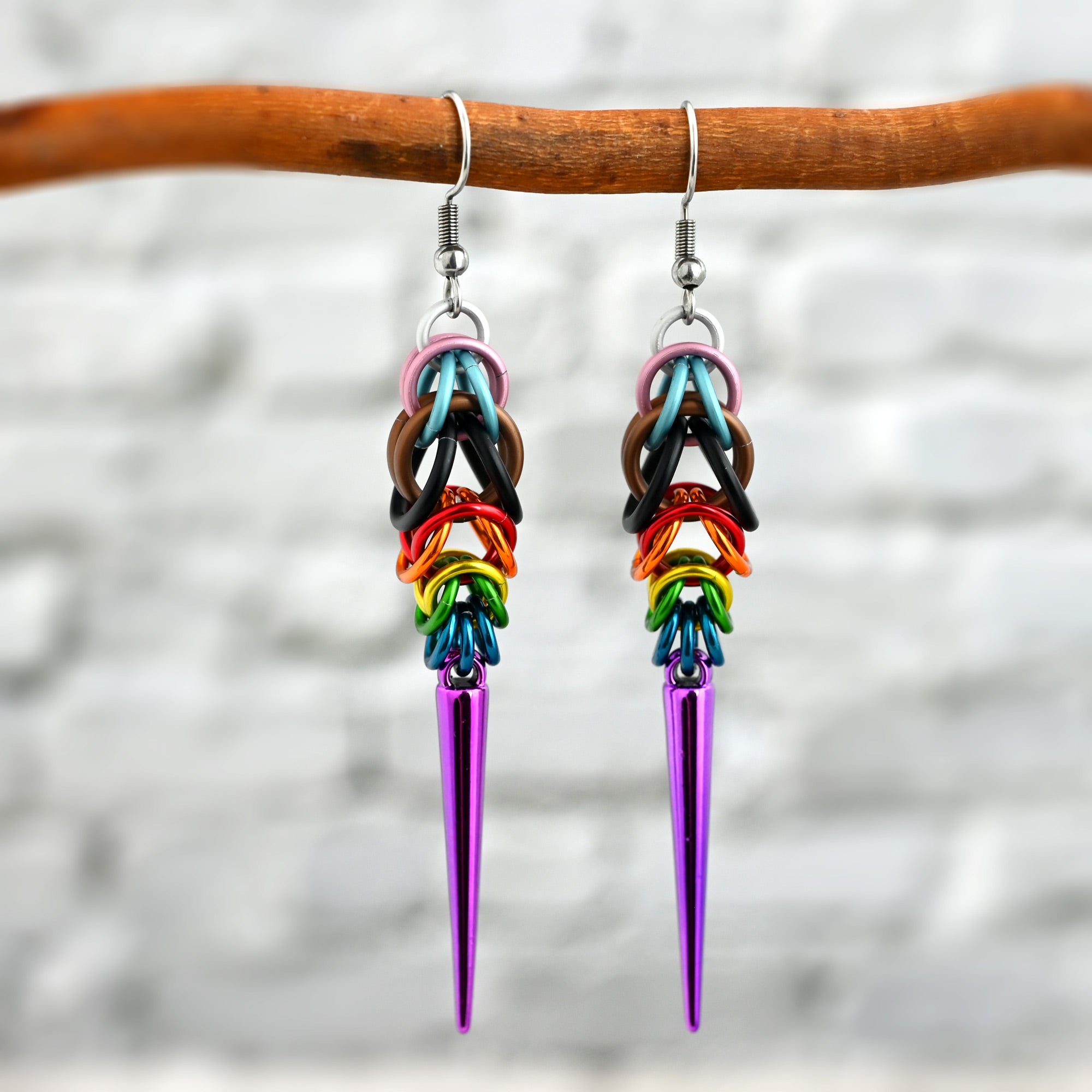 A pair of Progress Pride chainmaille spike earrings hanging from a natural wood branch. The earrings contain the 11 colors of the lgbtq progress pride flag, starting with white at the top. They are in an elongated teardrop shape with a long violet spike at the base.