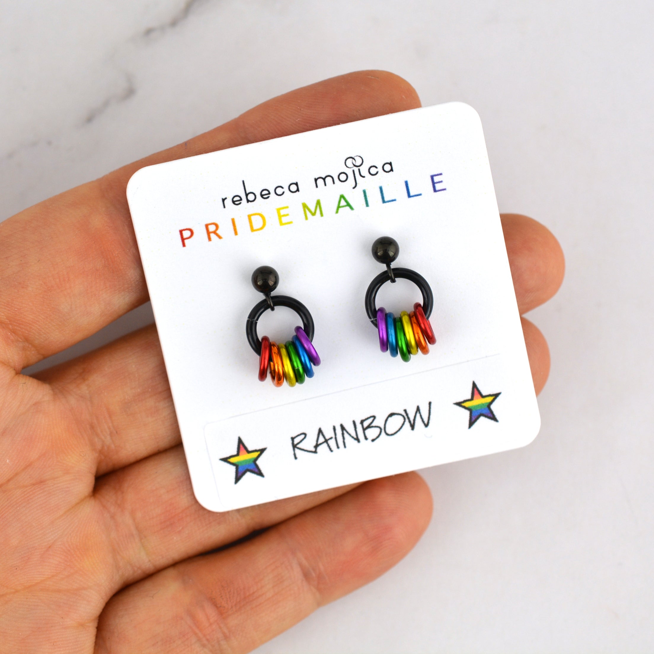 A small square white display card featuring two rainbow pride earrings is balanced on chainmaille artisan Rebeca Mojica’s fingertips. The card reads "rebeca mojica PRIDEMAILLE" and "RAINBOW" with stars.