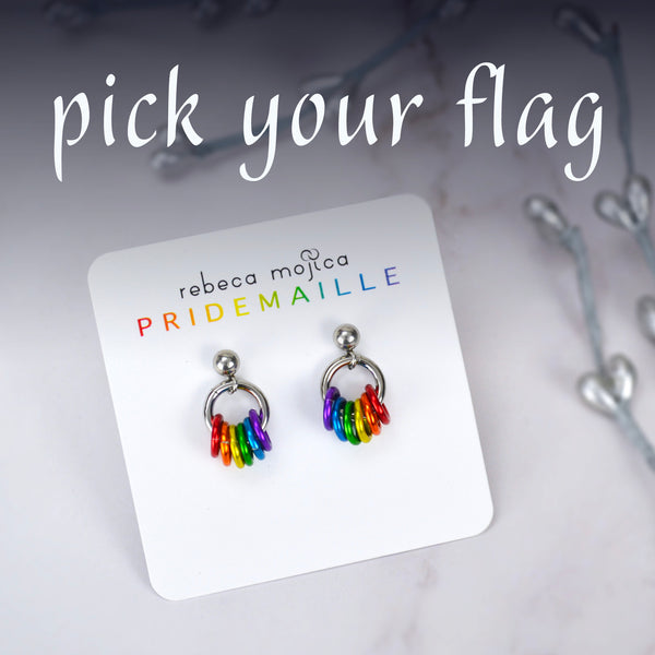  Tiny rainbow pride chainmaille earrings on a white earring card. There is large text at the top of the image that reads pick your flag. The earrings are a stud with a shiny 4mm ball. A small steel ring hangs from the ball. Six smaller colorful rings hang from the steel ring in the colors of the rainbow pride flag: red, orange, yellow, green, blue and violet. The earring card has the text Rebeca Mojica PRIDEMAILLE at the top.