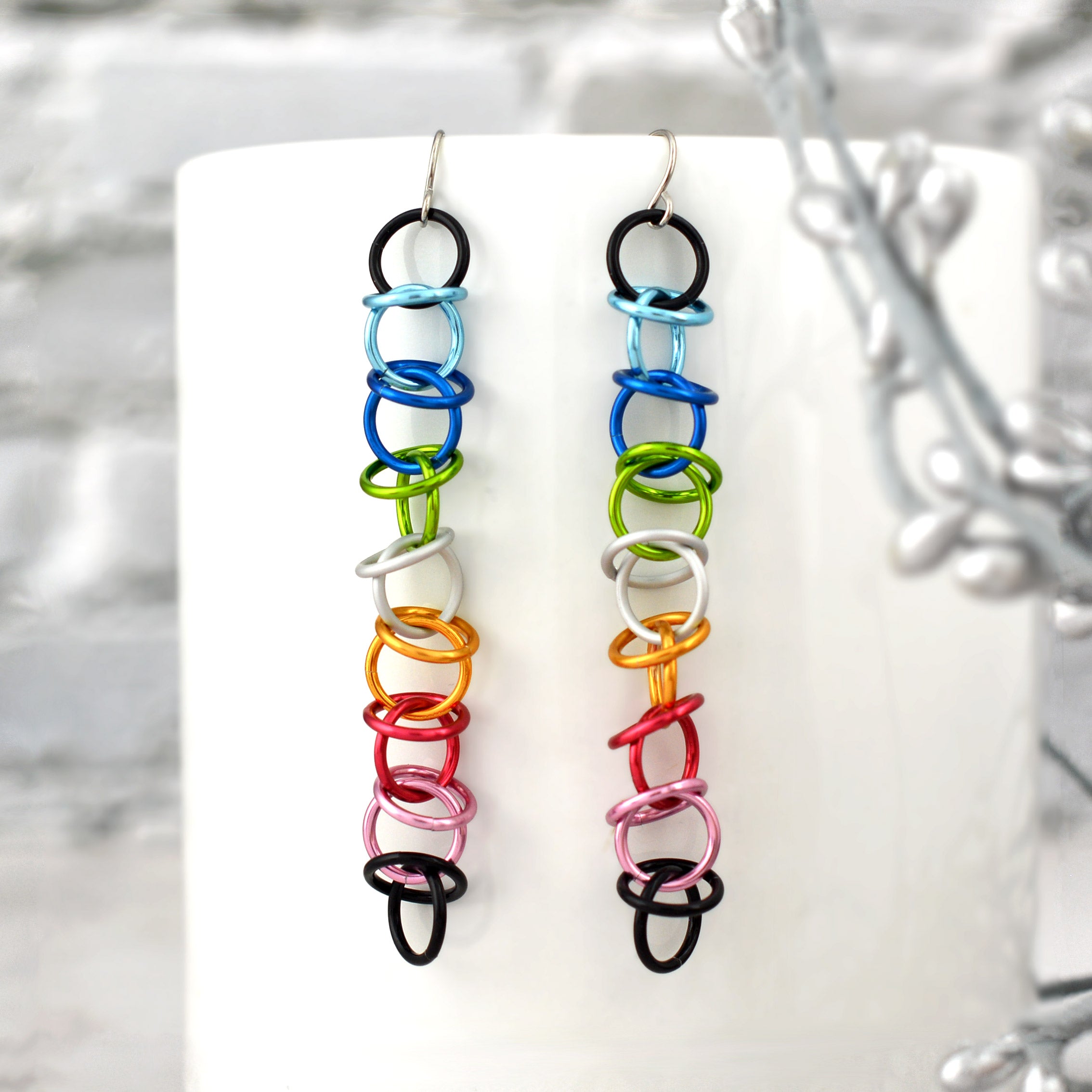 A pair of Queer Pride orbital earrings are showcased against a blurred setting of a smooth white mug and a touch of silver foliage. The earrings are composed of interconnected, vibrant aluminum rings in the Queer Pride flag colors: black, light blue, blue, bright green, white, gold, dark pink, pink, and black. Simple surgical steel earwires complete the design, highlighting their elegant dangle and vibrant colors.
